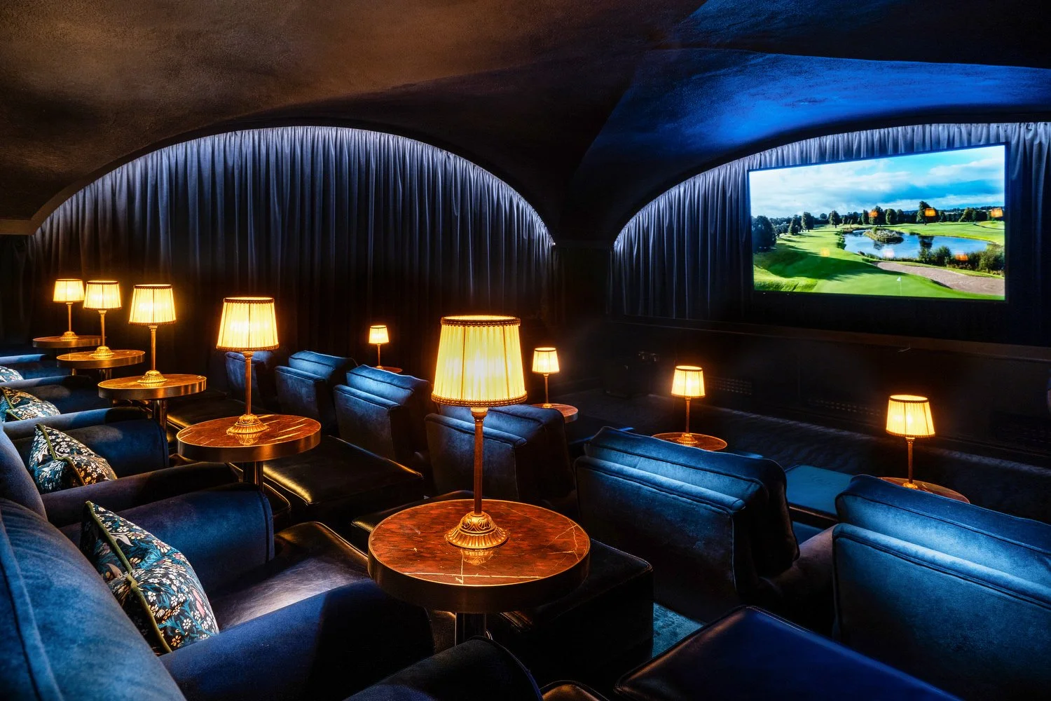 A luxurious lounge with blue velvet armchairs, wooden side tables with lamps, and a large screen showing a golf course landscape.