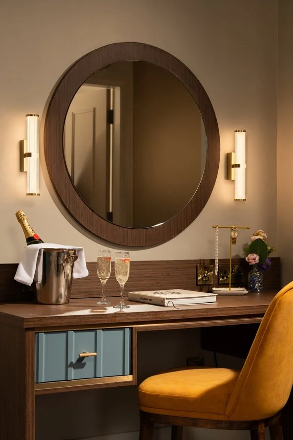 A wooden vanity with a round mirror, two wall-mounted lights, a yellow chair, a champagne bottle in an ice bucket, two glasses of champagne, a flower arrangement, and a white book.