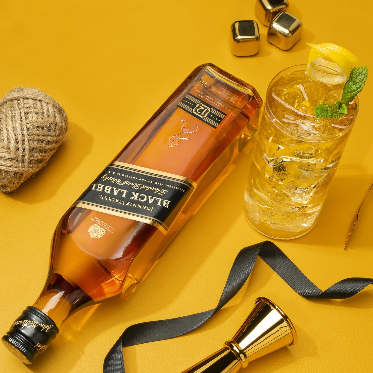A bottle of Johnnie Walker Black Label whiskey lying on a yellow surface next to a glass of ice filled with a drink garnished with a lemon wedge and mint, along with a jingle bell and a ball of yarn.