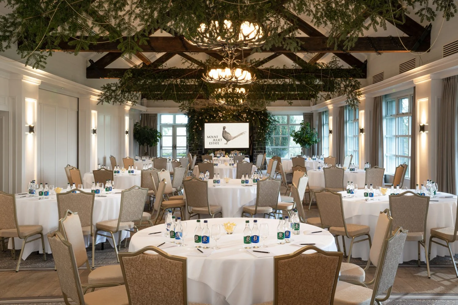 Elegant banquet hall decorated for a formal event with round tables, beige chairs, and bottled water. Greenery and a large screen displaying 'Mount Juliet Estate' are visible in the background, with large windows letting in natural light.