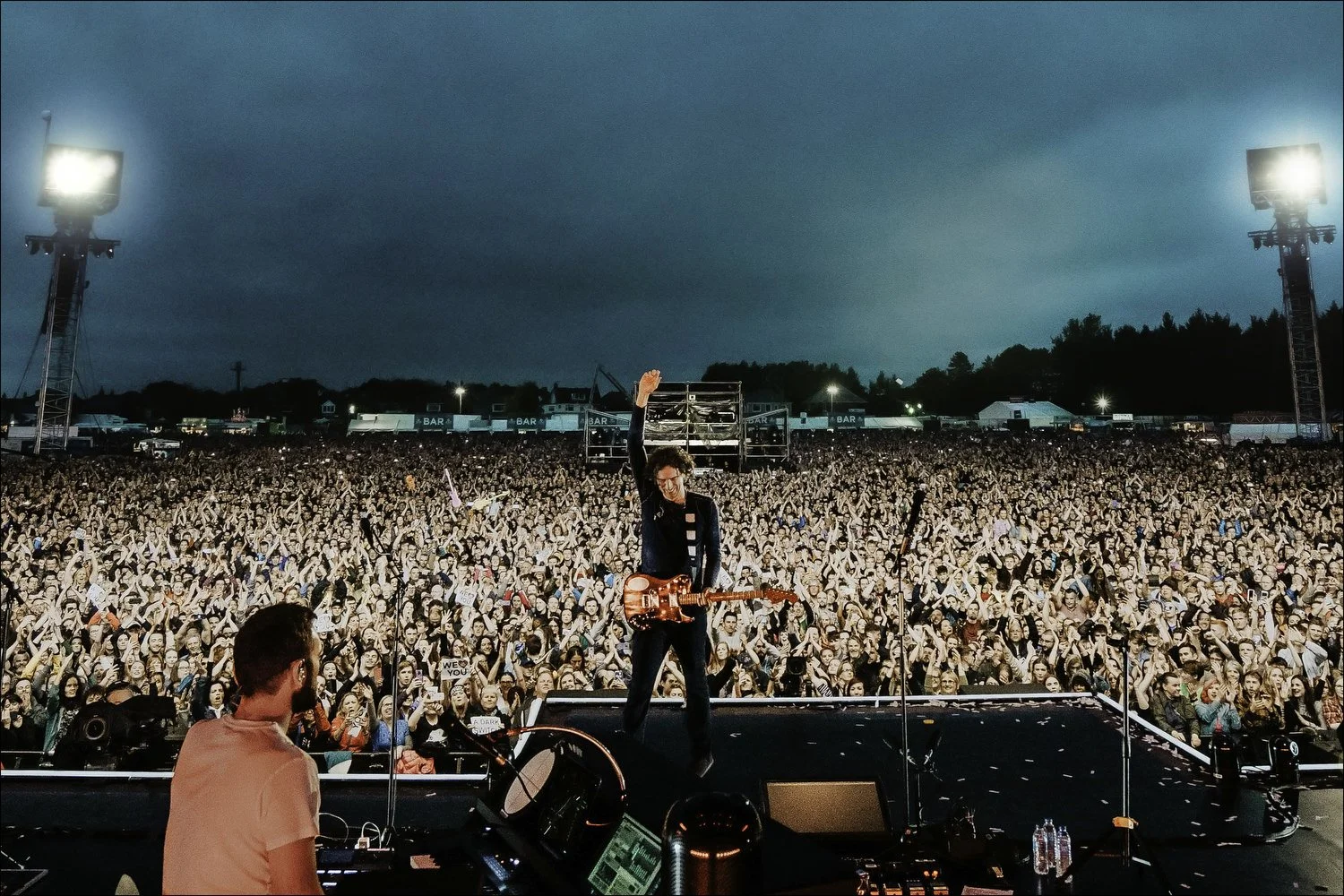 Performer on stage with guitar raising arm, large crowd at outdoor concert under darkening sky, concert lighting