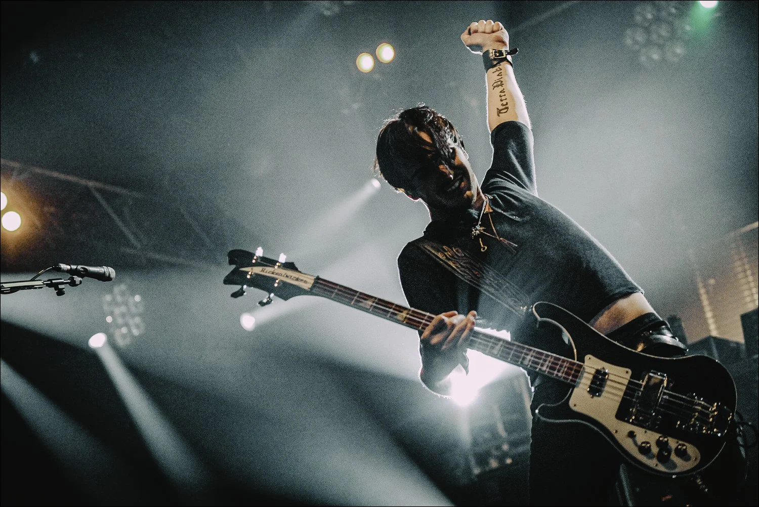 A musician performs on stage under green and white lights, playing an electric guitar and raising one hand in the air, with the background slightly blurred.