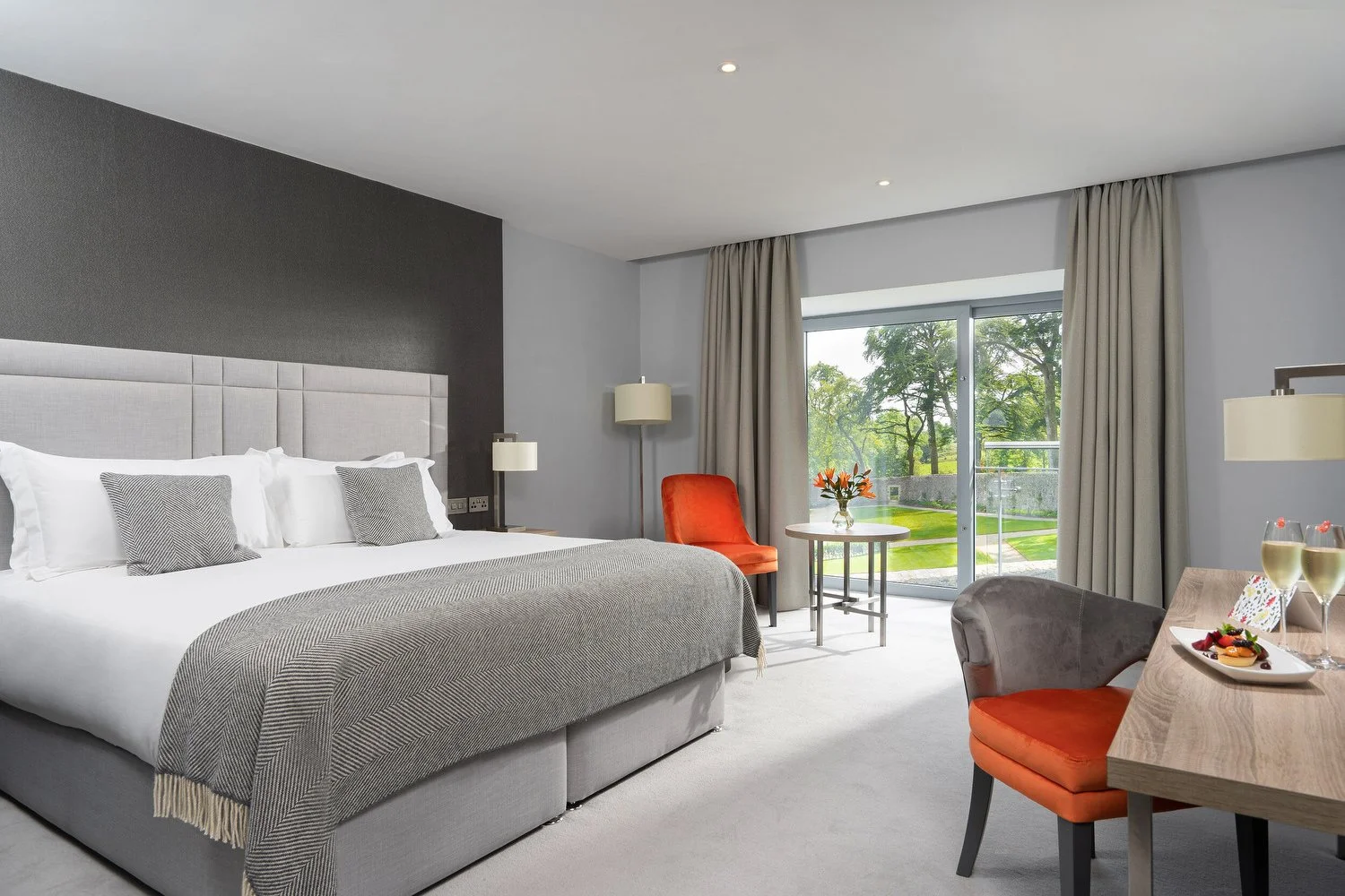 Hotel room with a large bed, white and gray bedding, two pillows, bedside lamps, a sliding glass door with green outdoor scenery, a small round table with a flower arrangement, colorful chairs, and a wooden desk with two glasses of champagne and a pl