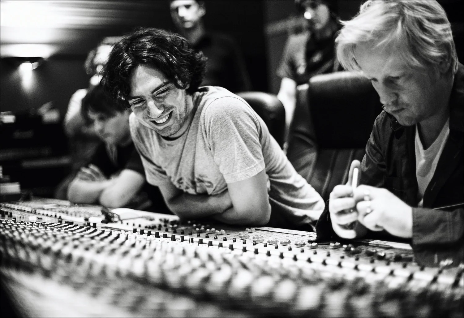 A group of men working in a music recording studio, some seated at a mixing console and others standing in the background.