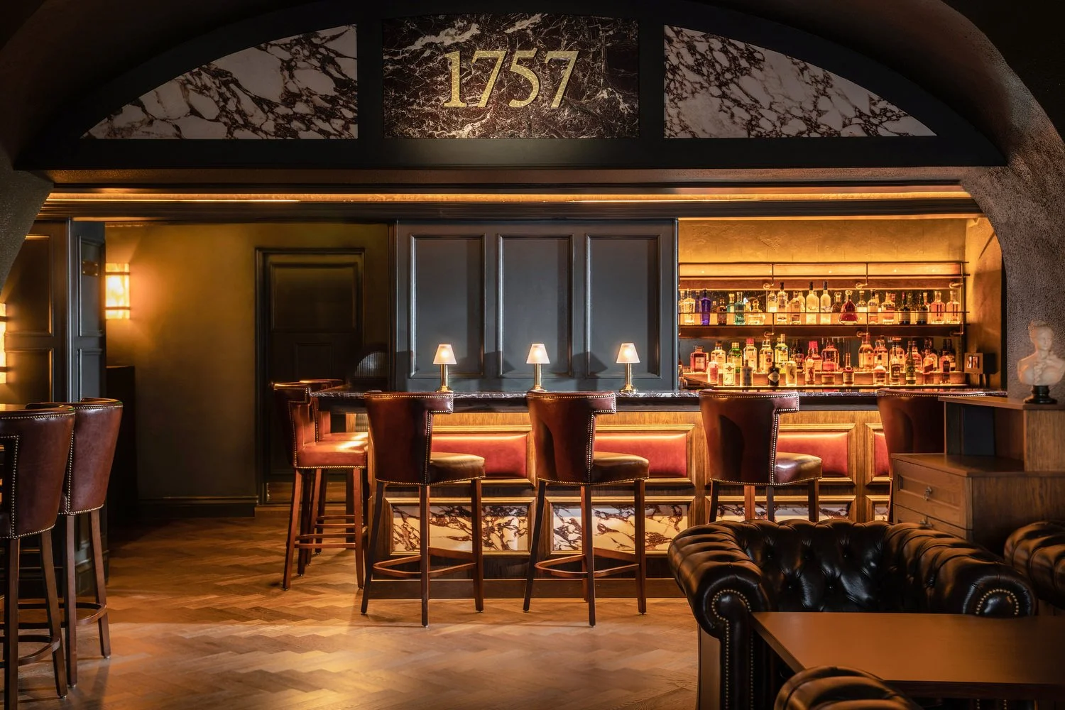 A stylish bar interior with dark wood accents, marble detailing, and warm lighting. A row of bar stools with leather upholstery line the marble-topped bar, which is stocked with various bottles of alcohol on illuminated shelves. The decor includes a 
