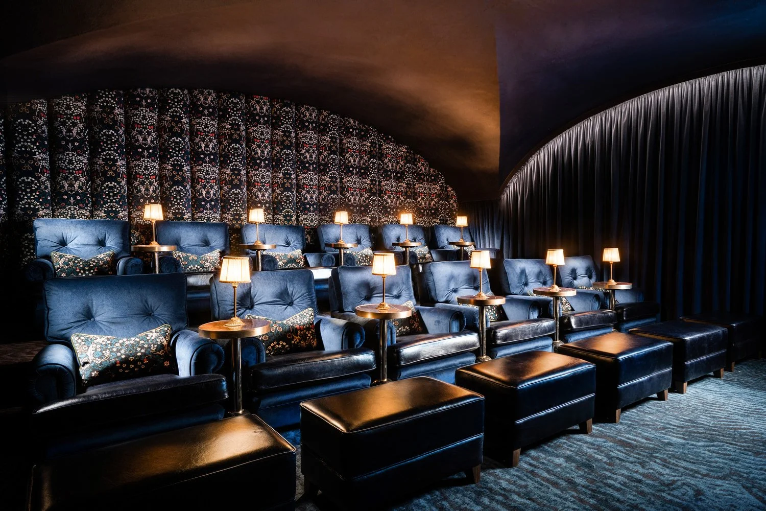 A dimly lit private theater with blue velvet seats, small table lamps, and dark patterned wallpaper.