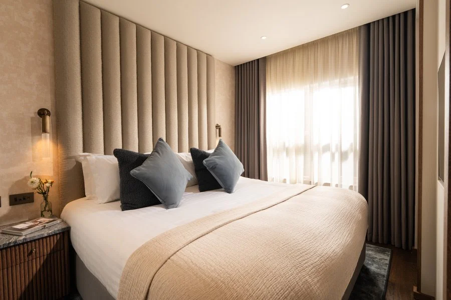 A modern bedroom with a large bed featuring beige and dark gray pillows, a padded beige headboard, a side table with a vase of flowers and magazines, and a window with sheer white curtains and dark drapes.