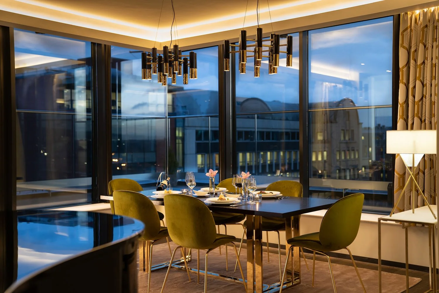 Elegant dining room with a black table set for six, surrounded by green chairs, large windows with city view, modern chandeliers, and a side lamp.
