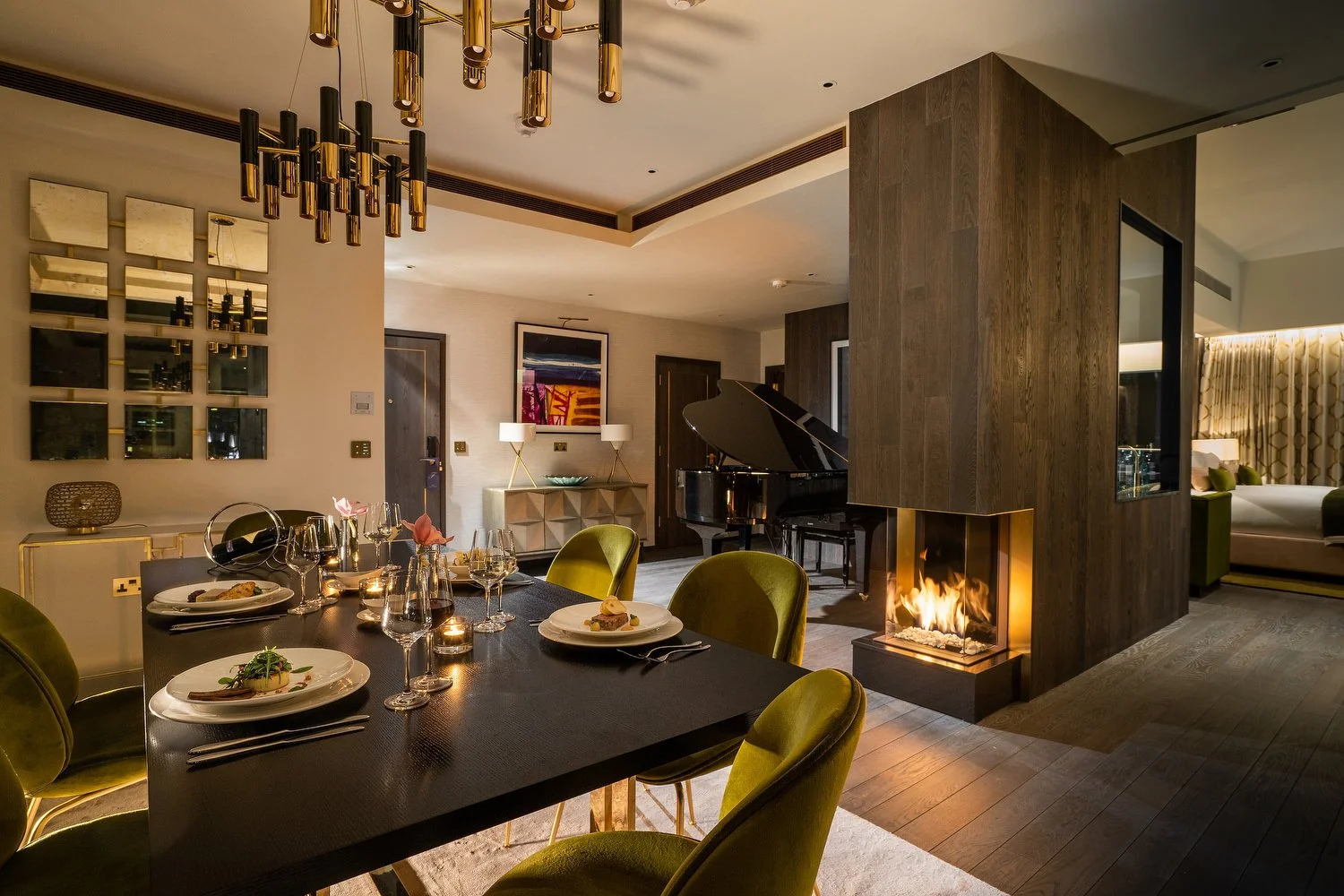 Elegant dining room with a black table set for a meal, surrounded by green velvet chairs, a modern fireplace, and a grand piano in the background.