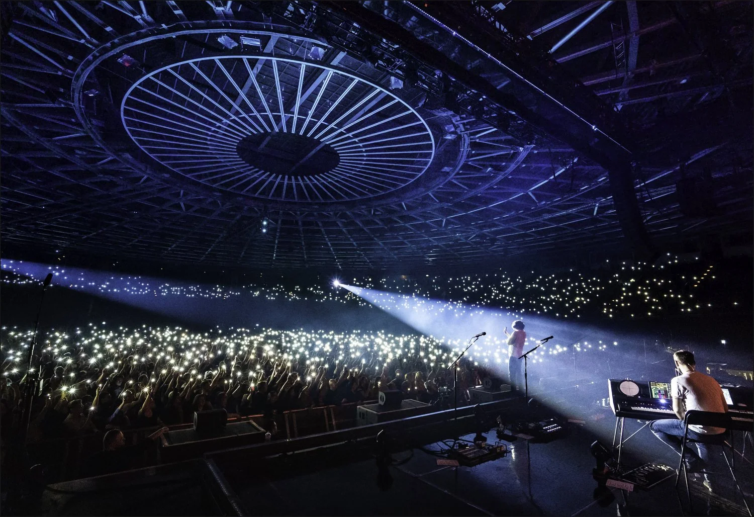 A concert with a performer on stage singing into a microphone, accompanied by a DJ or musician on a keyboard, in a large venue with a circular ceiling and a crowd holding up lights.