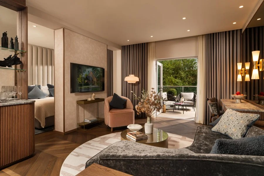 Modern living room with a view of a balcony and bedroom, featuring a dark gray sofa, pink armchair, coffee table with decorative items, wall-mounted TV, and large windows with curtains.