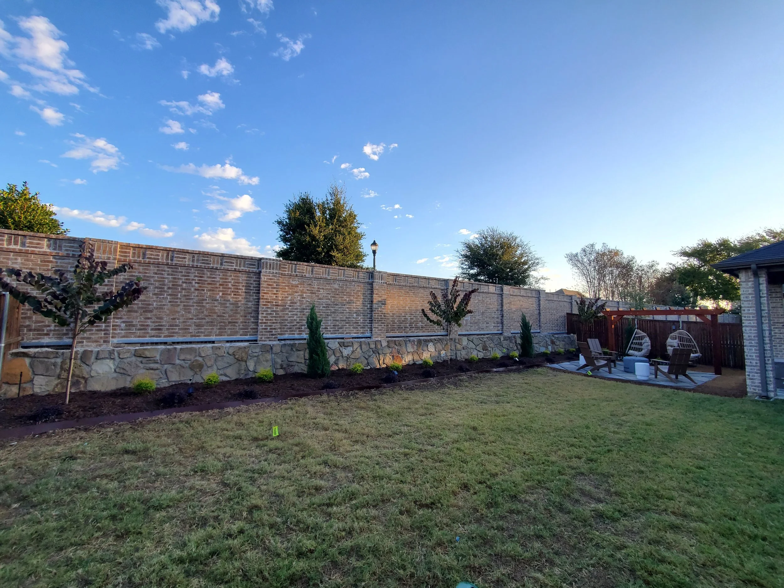A wide perspective of a backyard landscaping project installed by Designs of Eden, LLC, incorporating a newly installed flower bed, a firepit area with seating around and a pergola with two swinging chairs mounted securely.
