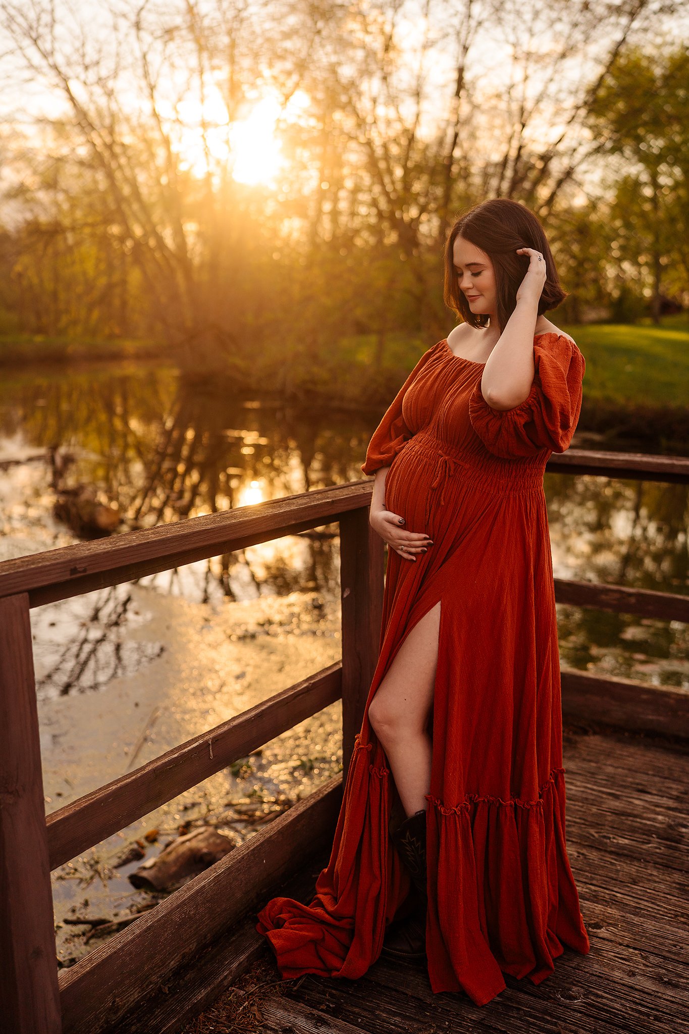 A pregnant woman in a flowing, off-the-shoulder rust-colored dress standing in a green field at sunset, smiling and looking to the side.
