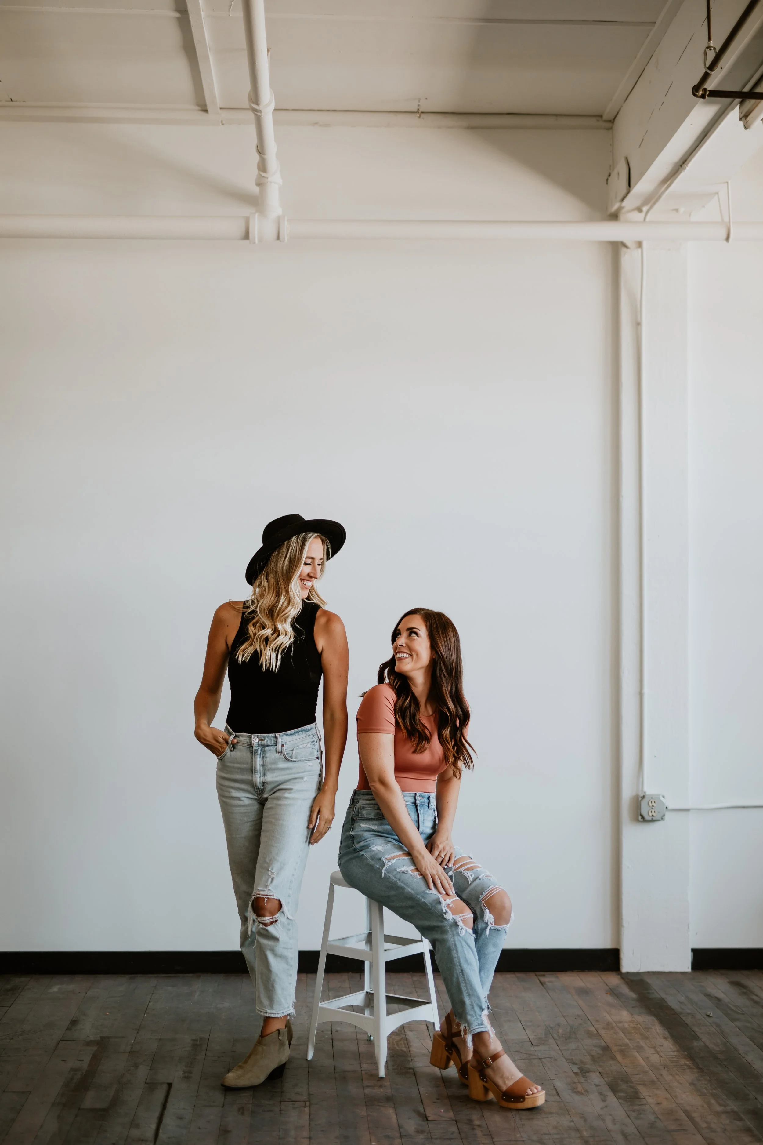 Two young women are in a minimalistic room with a white wall, hardwood floors, and exposed ceiling pipes. One woman with long blonde hair, wearing a black sleeveless top, ripped jeans, and beige ankle boots, is standing and smiling. The other woman, with long brown hair, wearing a pink T-shirt, ripped jeans, and brown high-heeled shoes, is seated on a white chair and looking up at the standing woman.