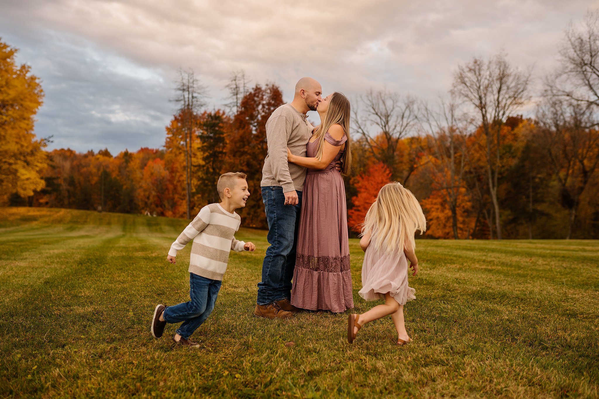 A family of five enjoying an autumn day outdoors, with trees in fall colors behind them. The couple is kissing, while the children are playing and running around on the grass.