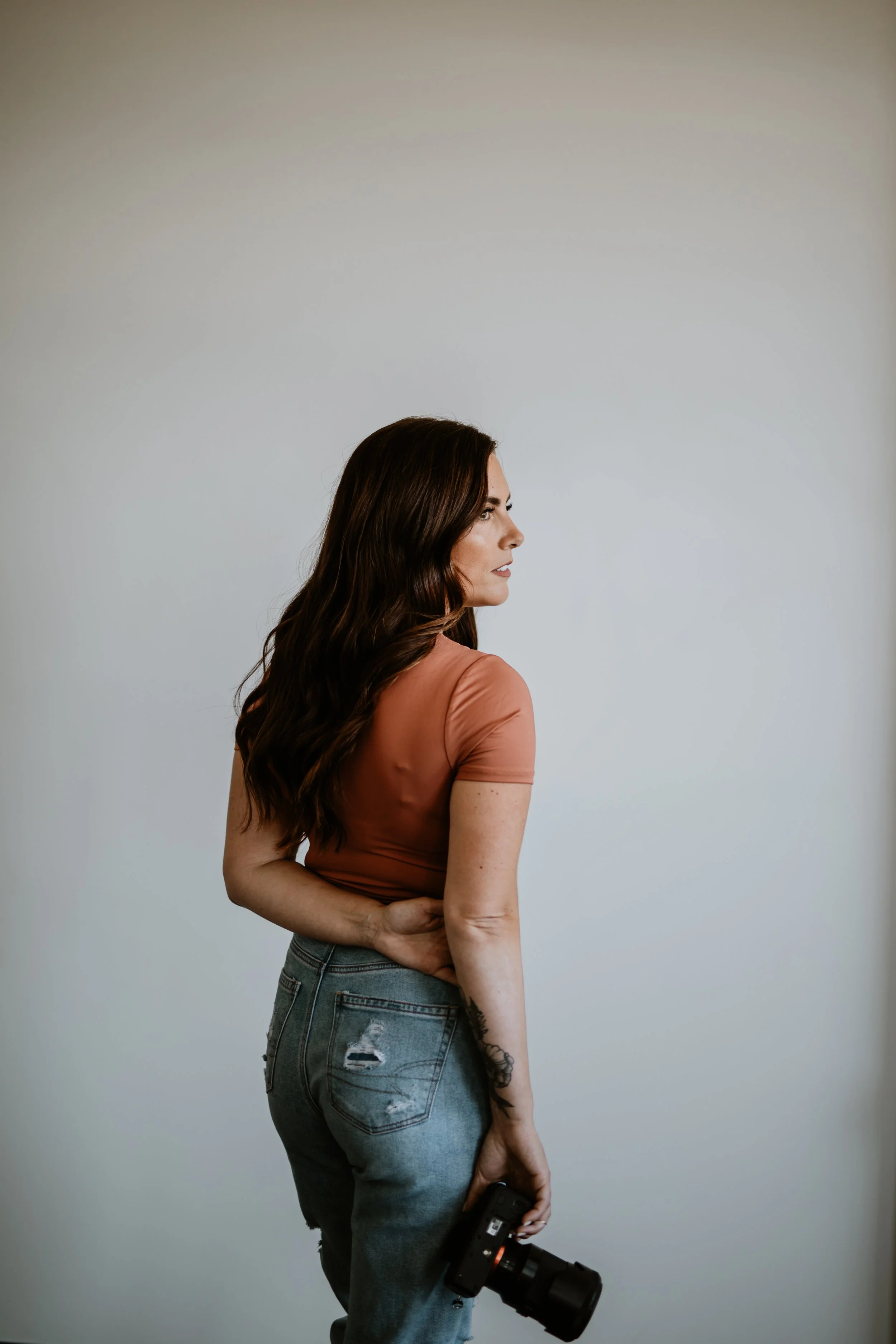 A woman with long wavy brown hair stands sideways against a plain white background. She is wearing a peach-colored t-shirt and ripped blue jeans, holding a camera in her right hand, with her left hand resting on her hip.