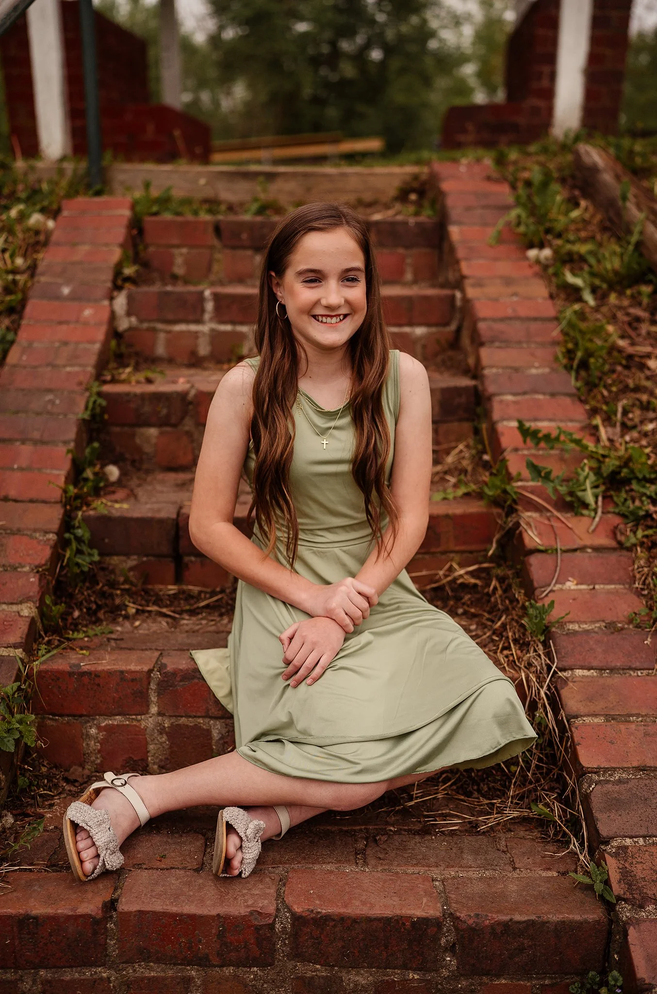 A young woman sitting on brick stairs outdoors, smiling, wearing a light green dress, earrings, and a necklace, with greenery and trees in the background.