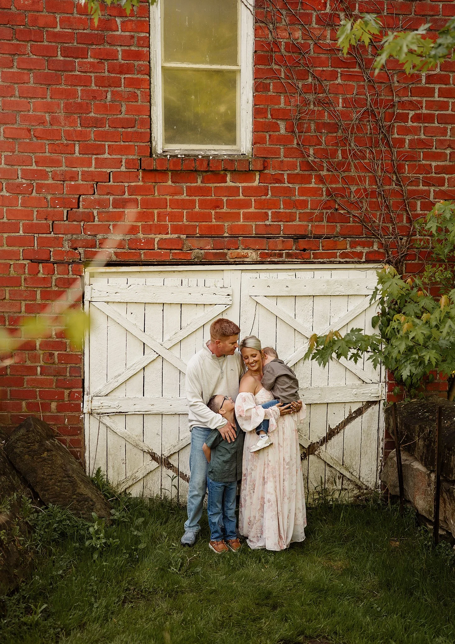 A family of four standing in front of a white wooden gate with a brick wall and window behind them. The adults are smiling, and the children are being held, one with glasses. There is green foliage and rocks in the foreground.