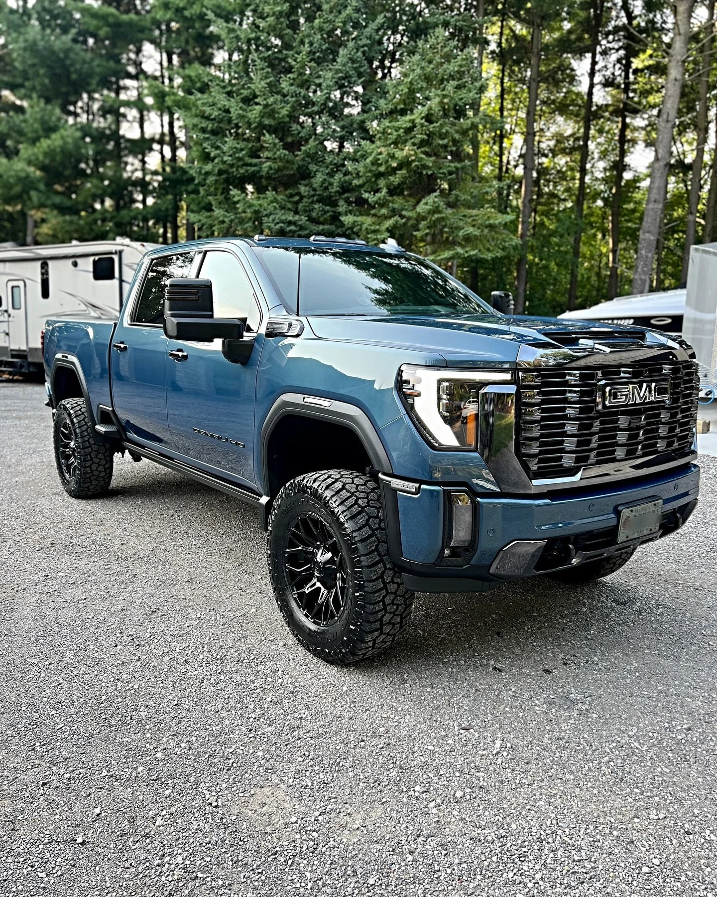 Blue GMC Sierra pickup truck with off-road tires parked on gravel, surrounded by trees and RVs.