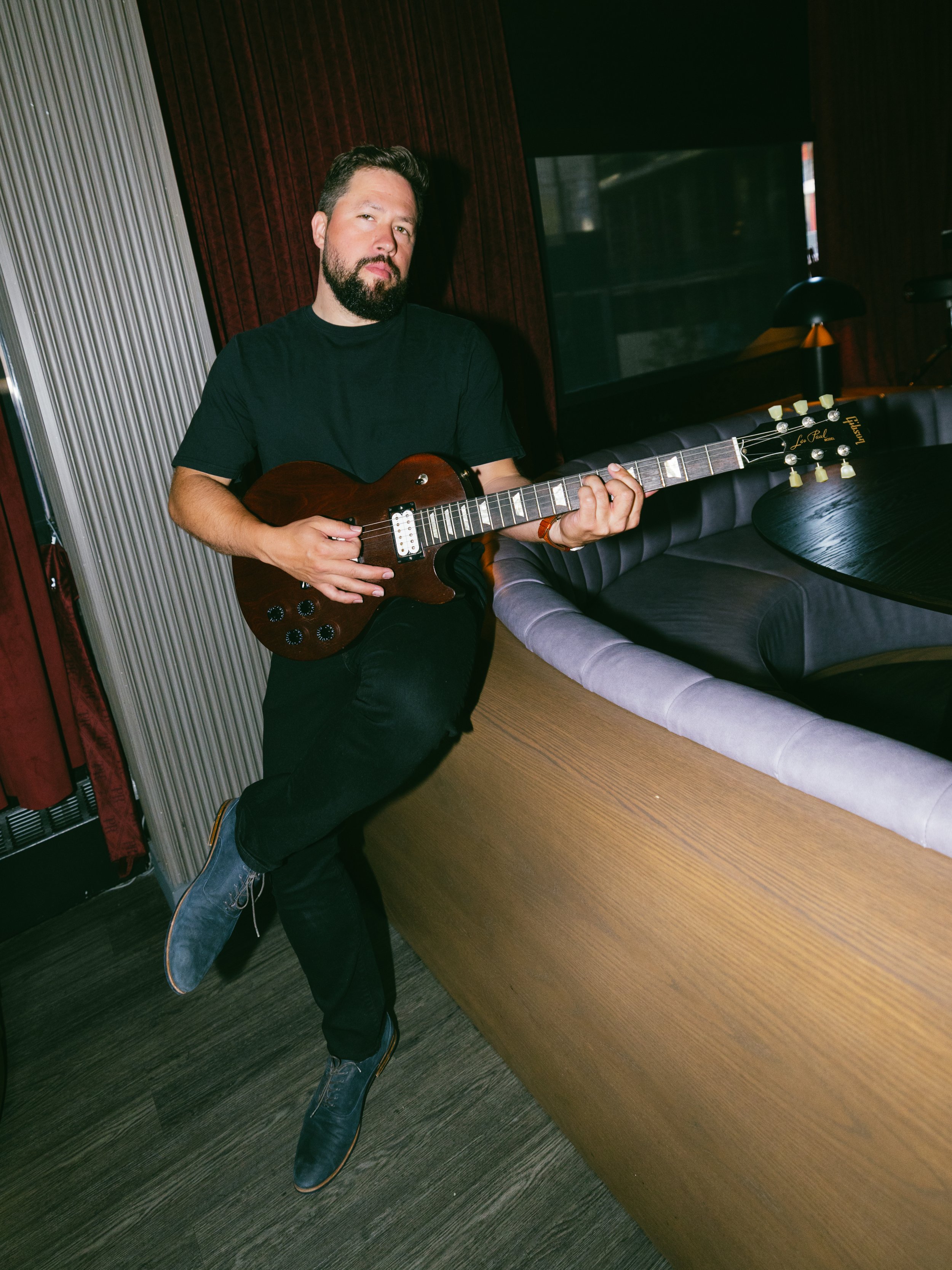 A man with a beard playing an electric guitar in a room with wood-paneled walls and a dark couch.