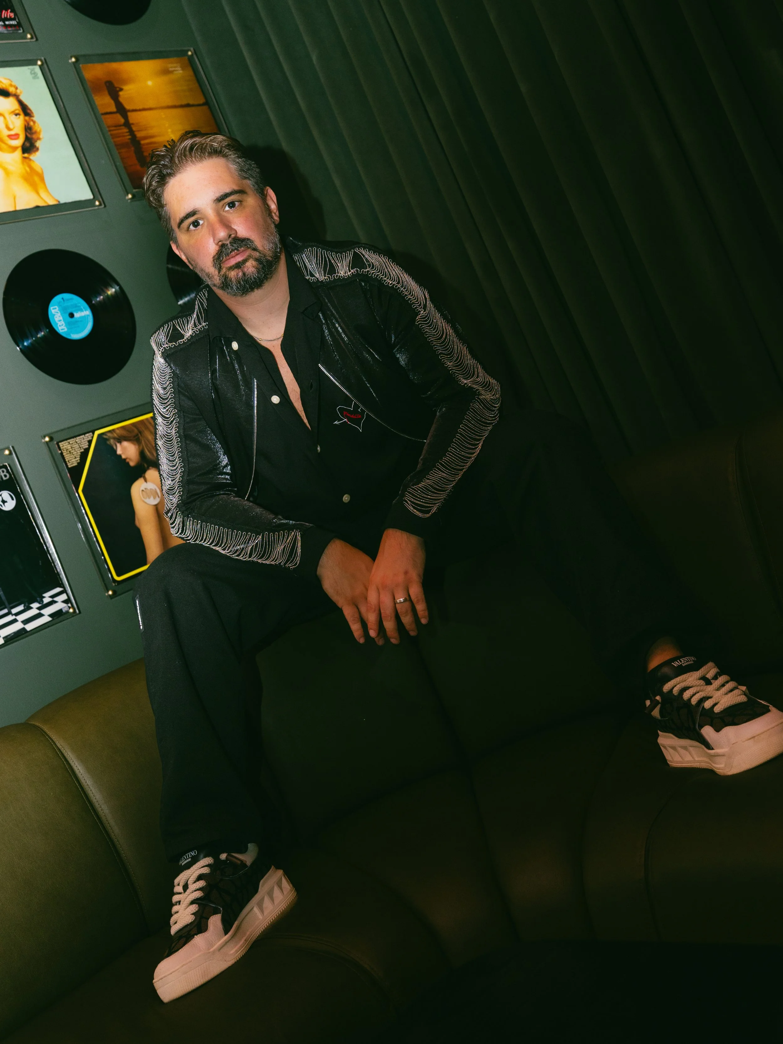 A man with a beard and gray hair sitting on a green couch, wearing a black jacket with white embroidered lines on the sleeves, a black shirt, and sneakers with beige soles. Behind him are dark green walls with framed pictures, one of a woman and another of a sunset over water, and a vinyl record on the wall.