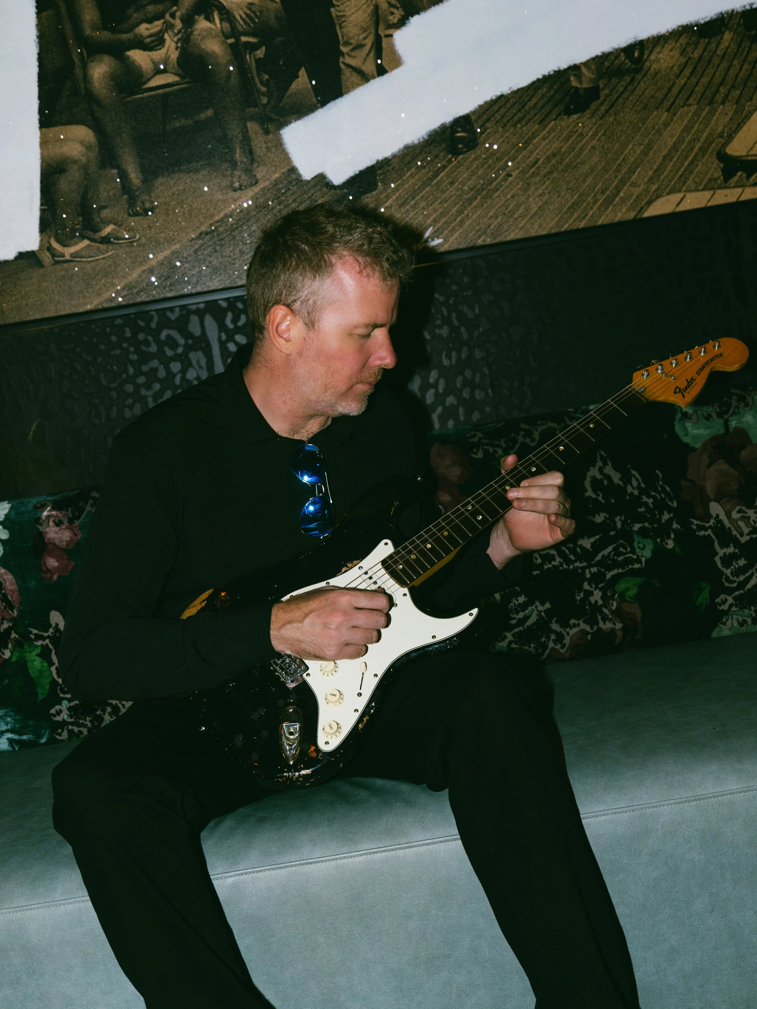 A man with short gray hair, wearing a black shirt, is sitting on a gray bench and playing an electric guitar with a black body and a white pickguard. In the background, there's a mural or painting on the wall depicting people sitting on a couch, with snow falling around them.