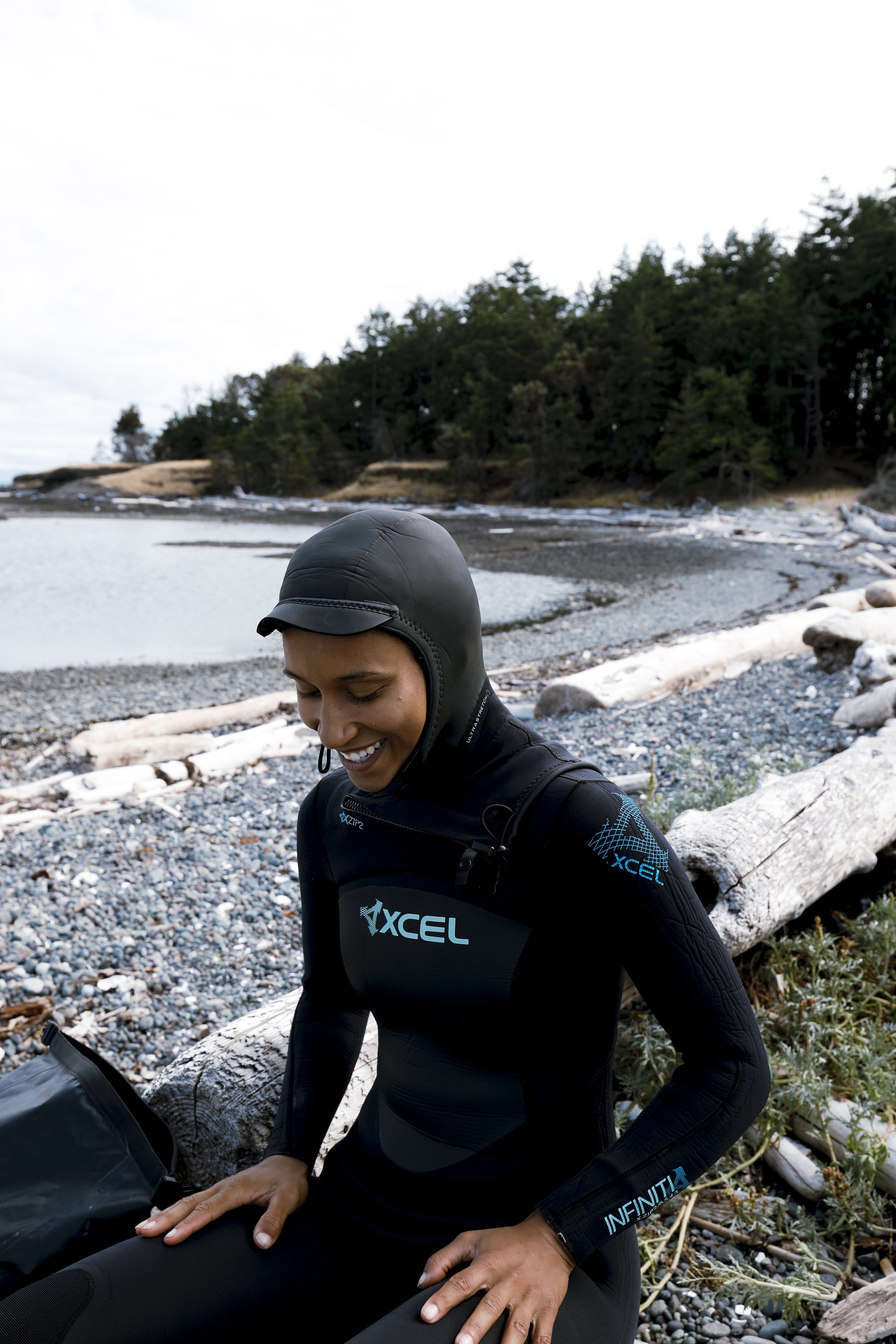 A woman wearing black wetsuit and helmet smiling on a rocky beach with driftwood and trees in the background.