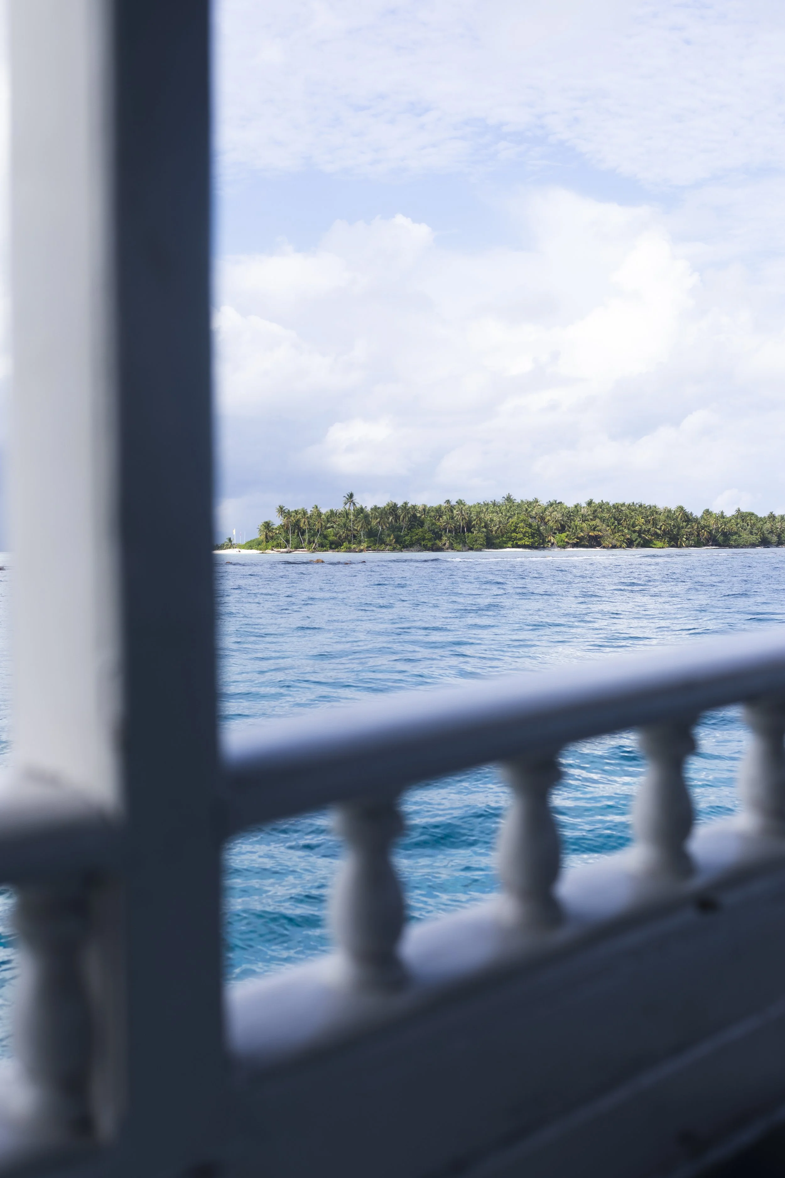 View of a tropical island with lush green trees seen through the window of a boat, with water in the foreground and partly cloudy sky above.