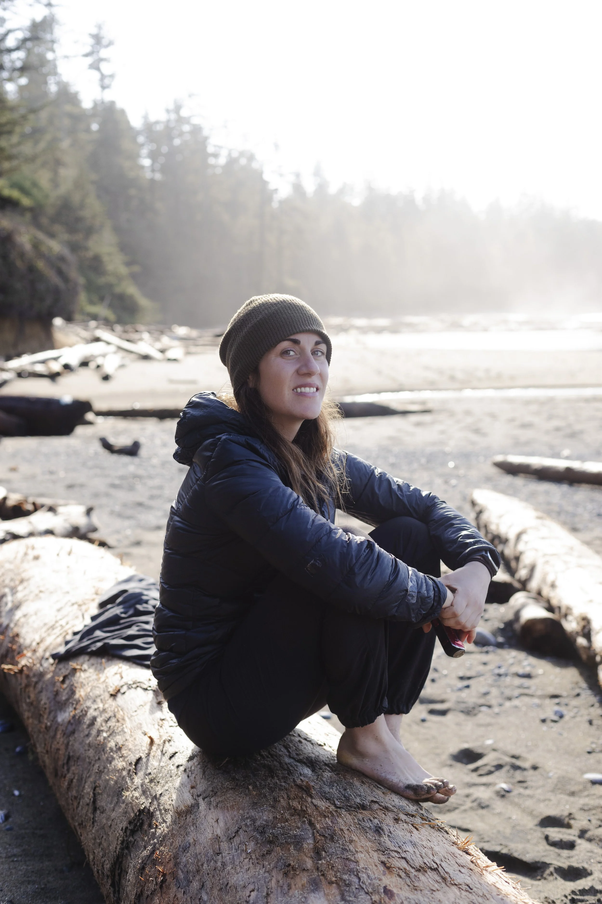 A woman sitting barefoot on a large driftwood log on a beach, wearing a dark jacket and knit hat, facing slightly to the right, with trees and ocean in the background.