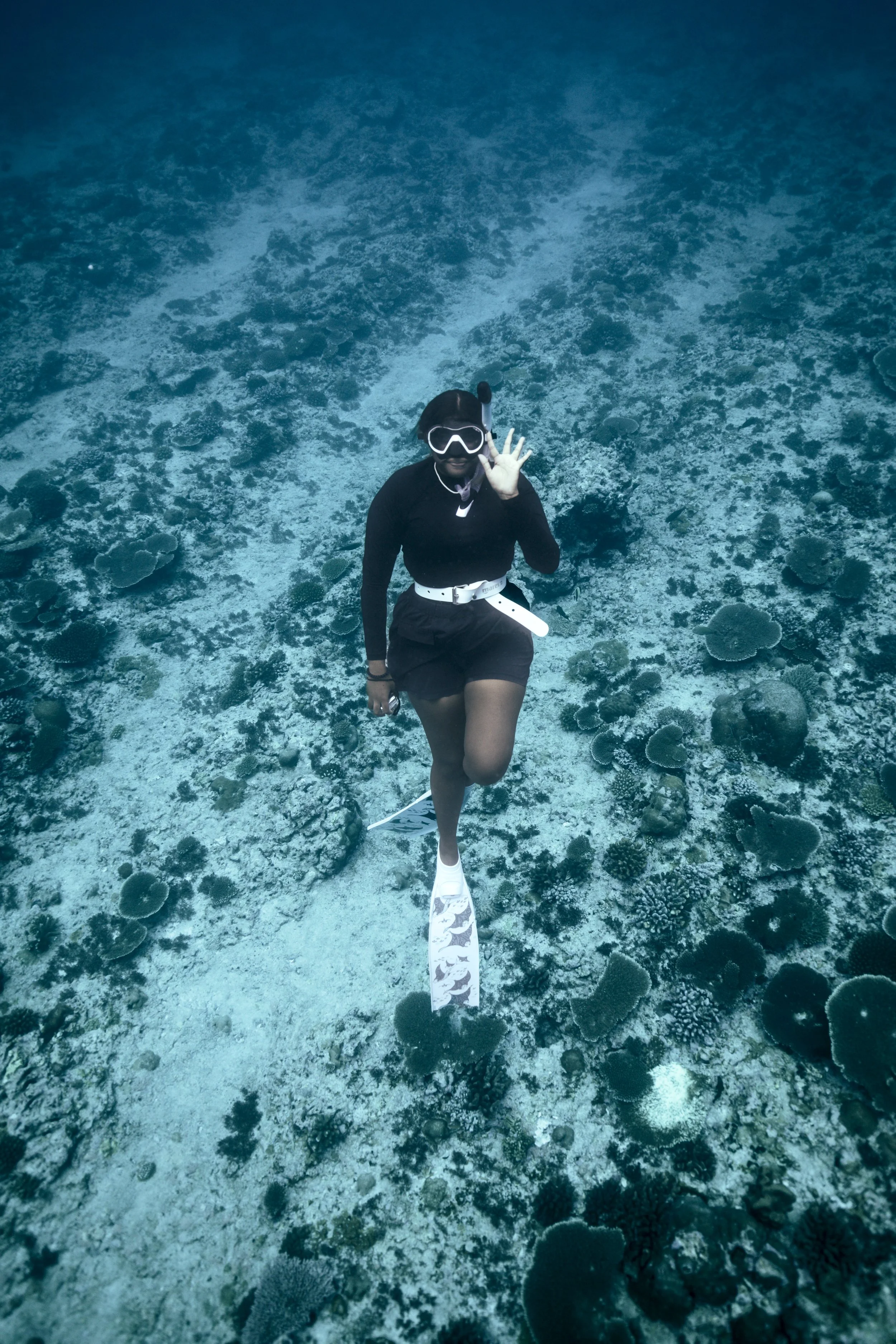 A person snorkeling underwater, waving and smiling, surrounded by coral and marine life.