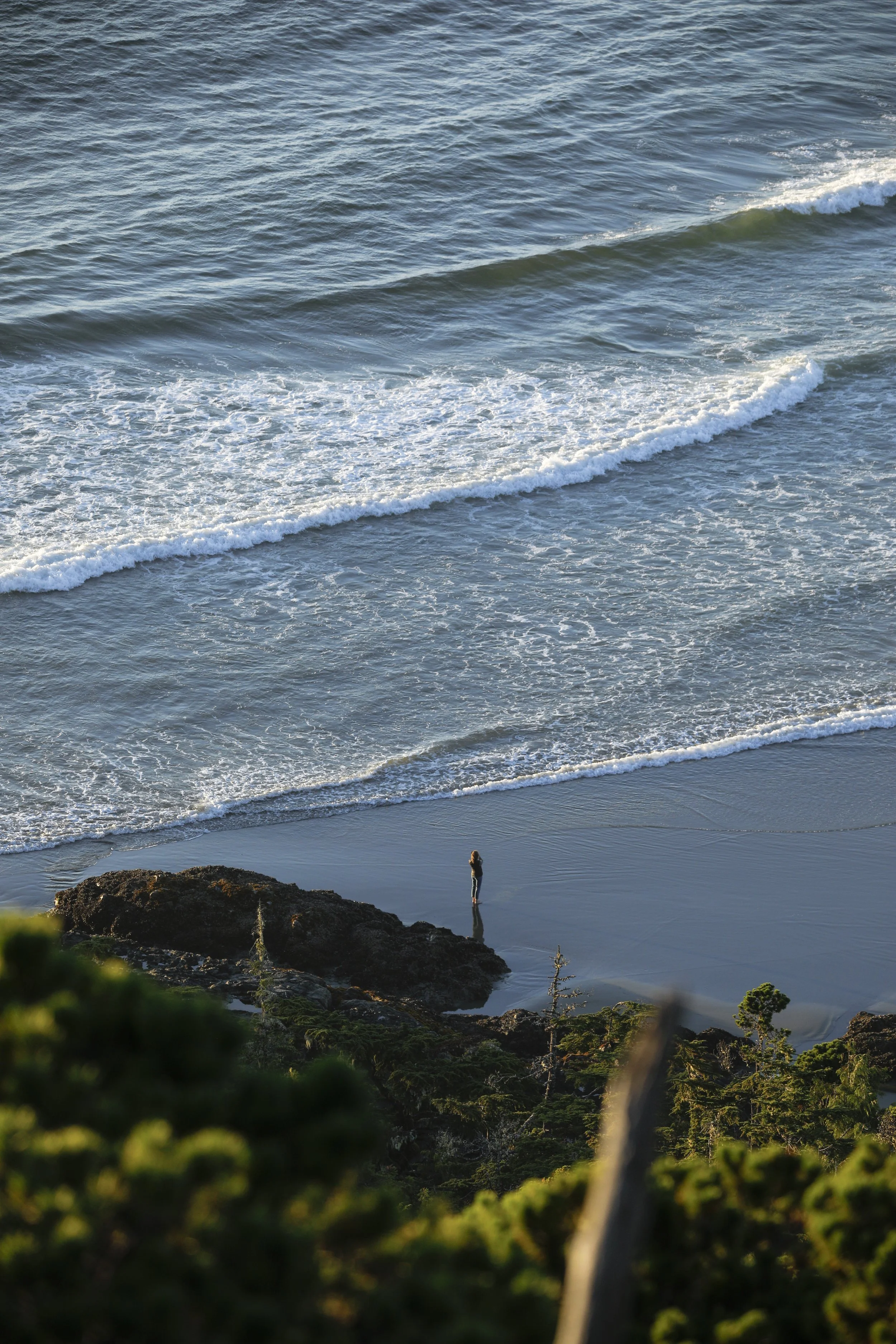 A person stands on a rocky outcrop at the beach, overlooking the ocean with small waves crashing onto the shore, in the late afternoon or early evening.