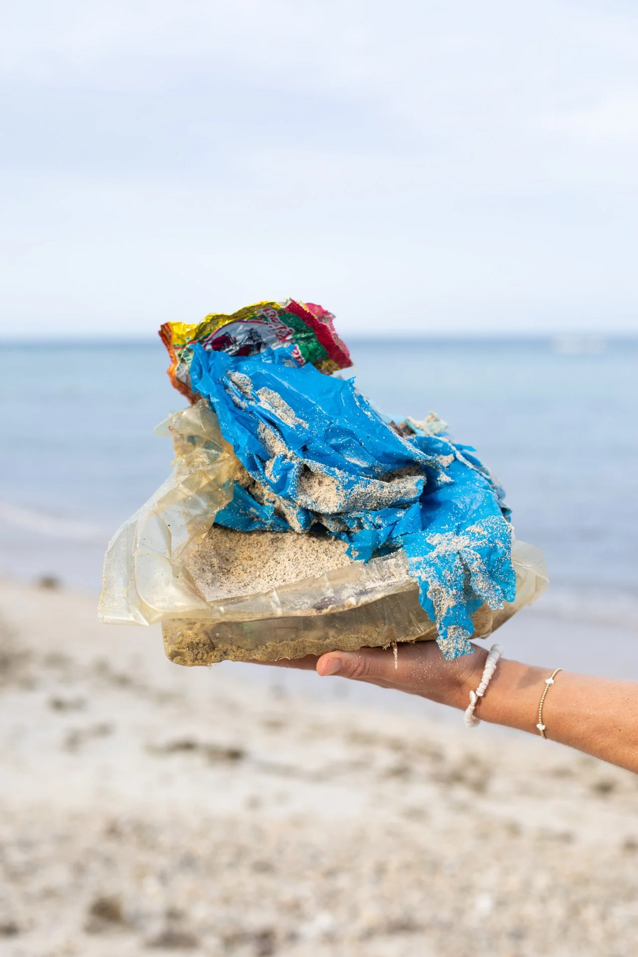 Person holding a large piece of trash, including plastic bags and debris, on the beach during daytime.