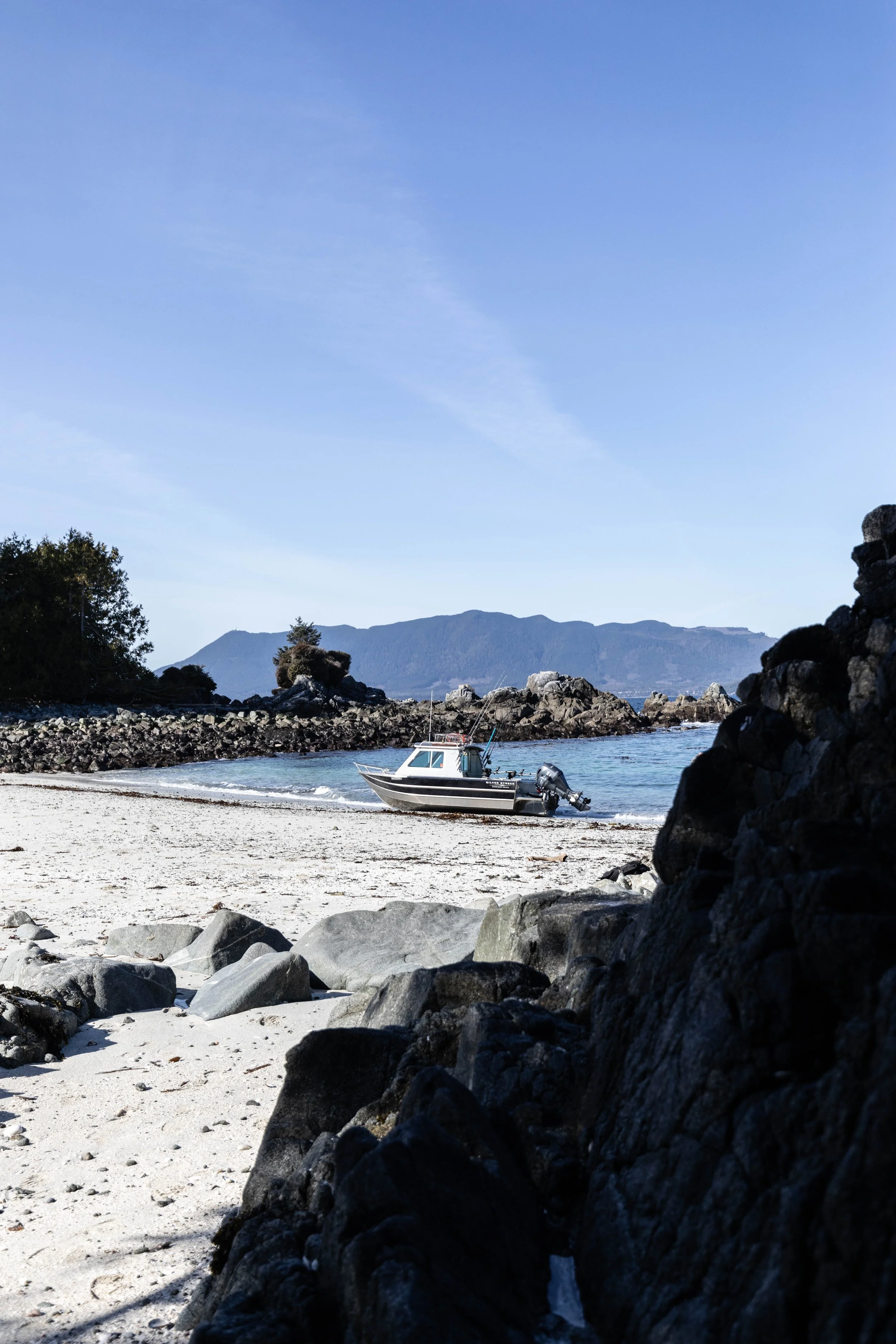 Boat resting on a sandy beach near rocky shoreline, with calm water, rocky islands, and a mountain range in the background under a clear blue sky.