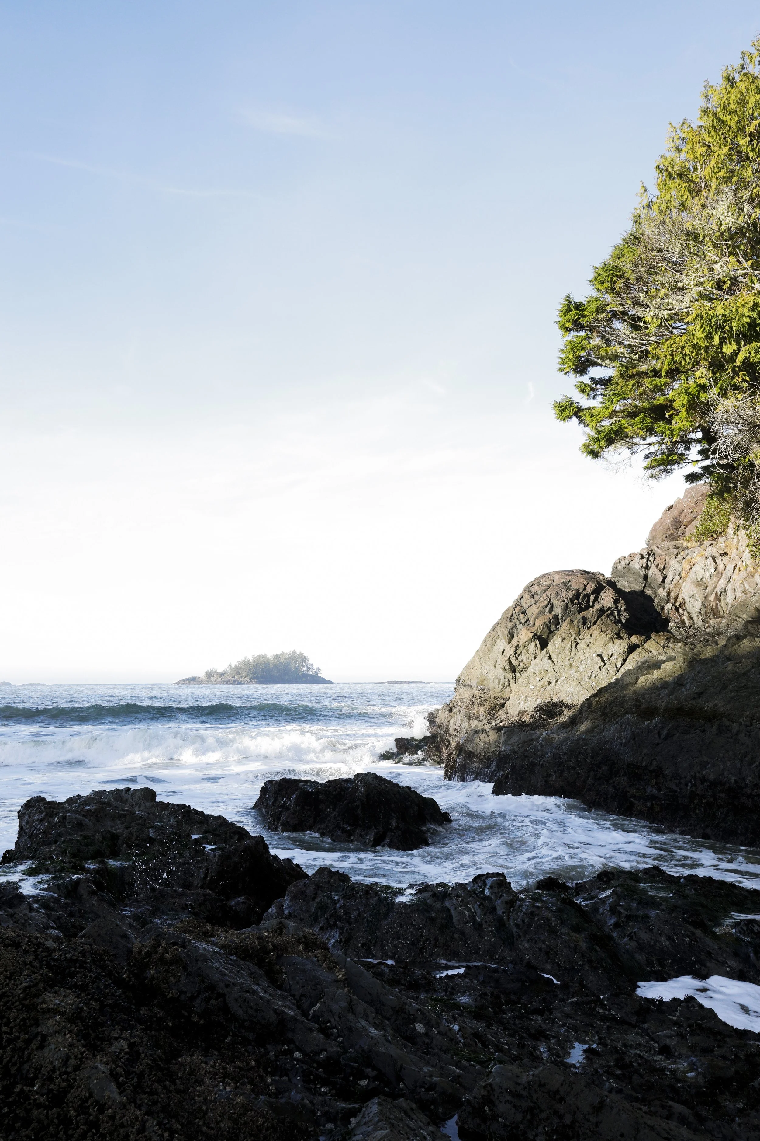 A rocky shoreline with waves crashing against dark rocks, a small island in the distance, and a partly cloudy sky.