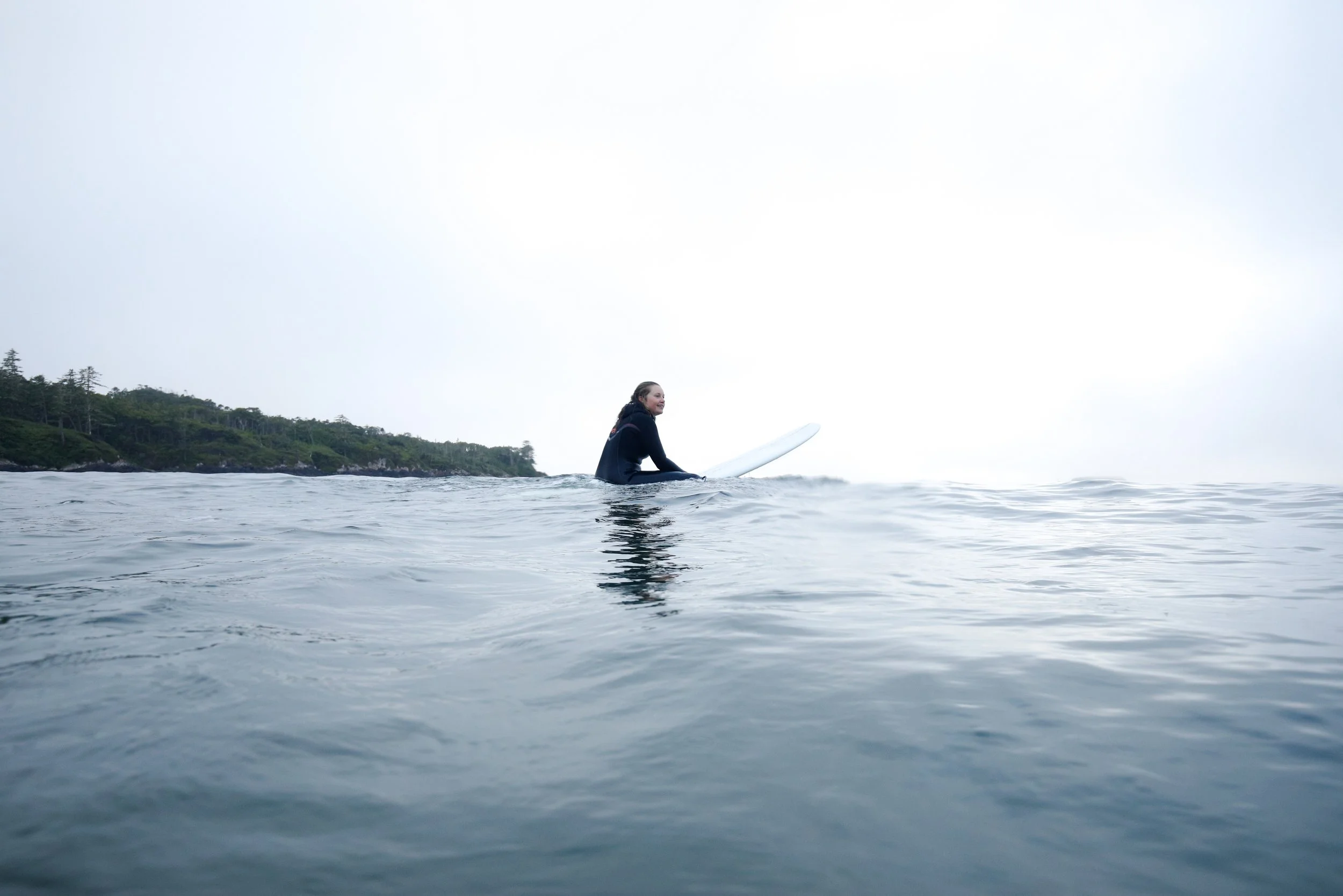 A person sitting on a surfboard in the ocean during a cloudy day, with a distant shoreline and trees in the background.