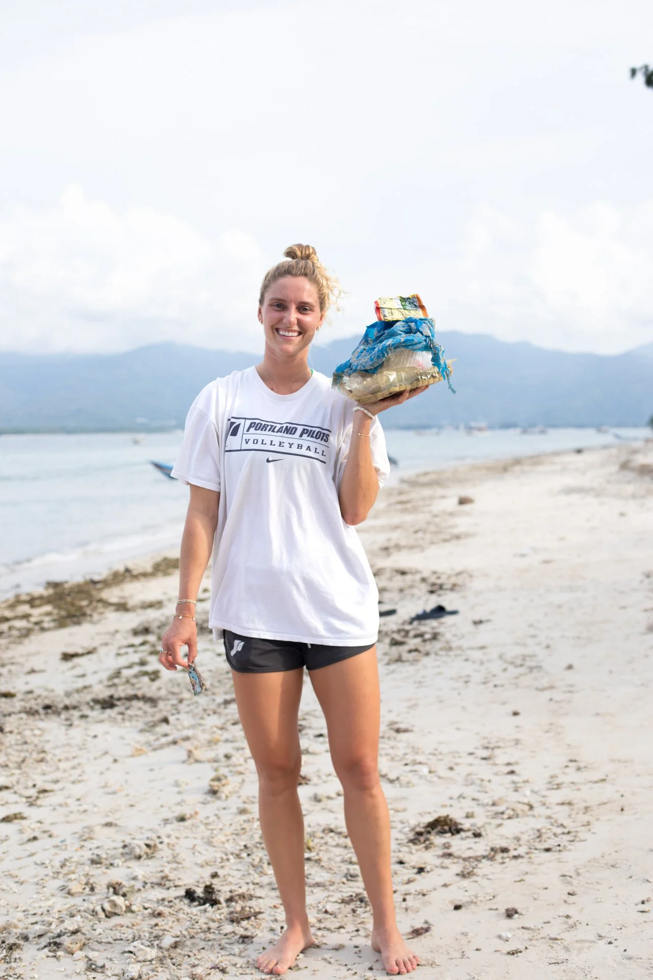 A young woman smiling at the camera while standing barefoot on a beach, holding a bag of collected trash, with a scenic background of water and mountains.
