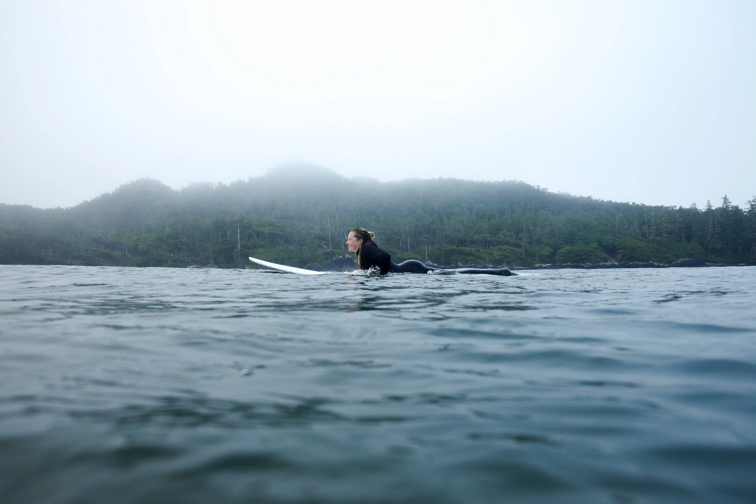 A woman in a black wetsuit paddling on a surfboard in calm water with a forested, foggy hillside in the background.