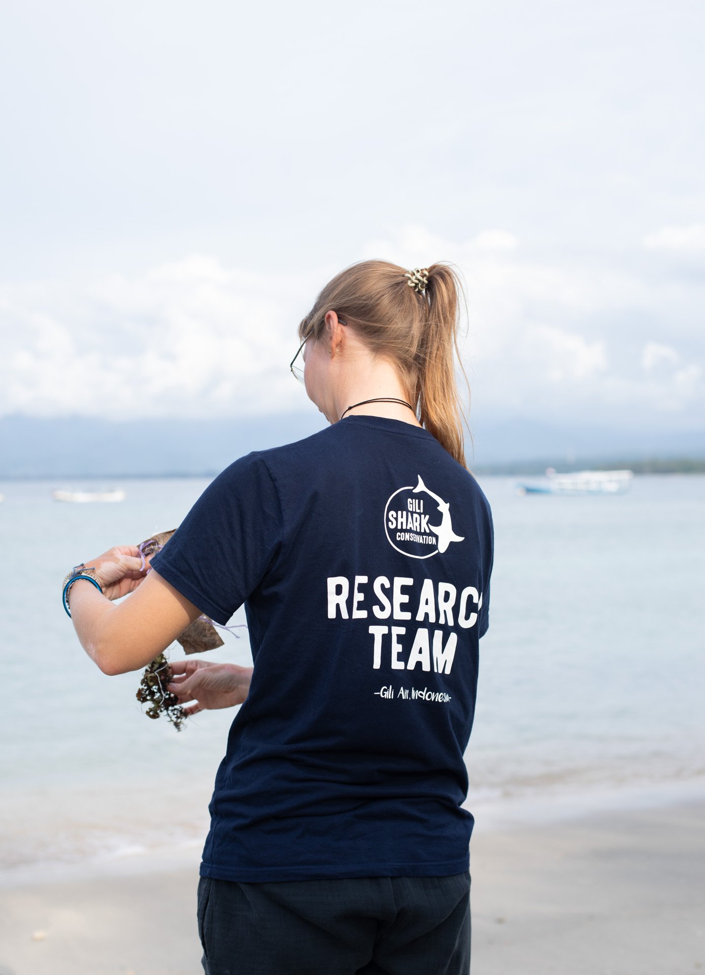 A woman with long blonde hair tied back, wearing glasses and a navy blue T-shirt with white text that reads "RESEARCH TEAM" and a logo for Gilli Shark Conservation, stands by the water at the beach holding a seaweed or marine sample.