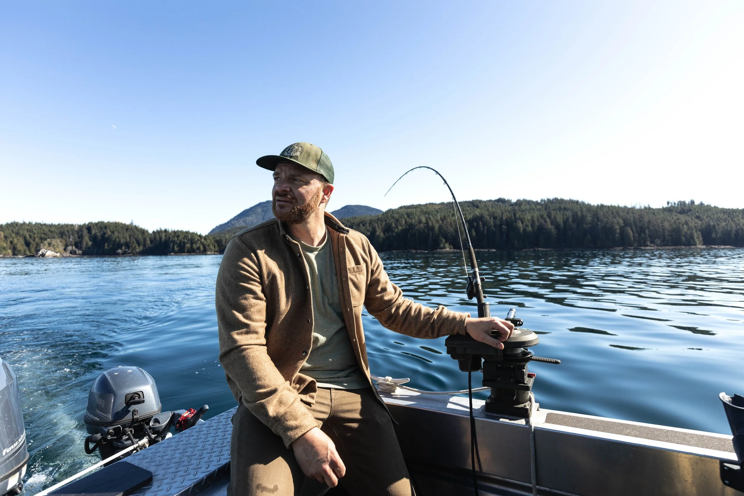 A man fishing from a boat on a calm lake with a forested shoreline and hills in the background.