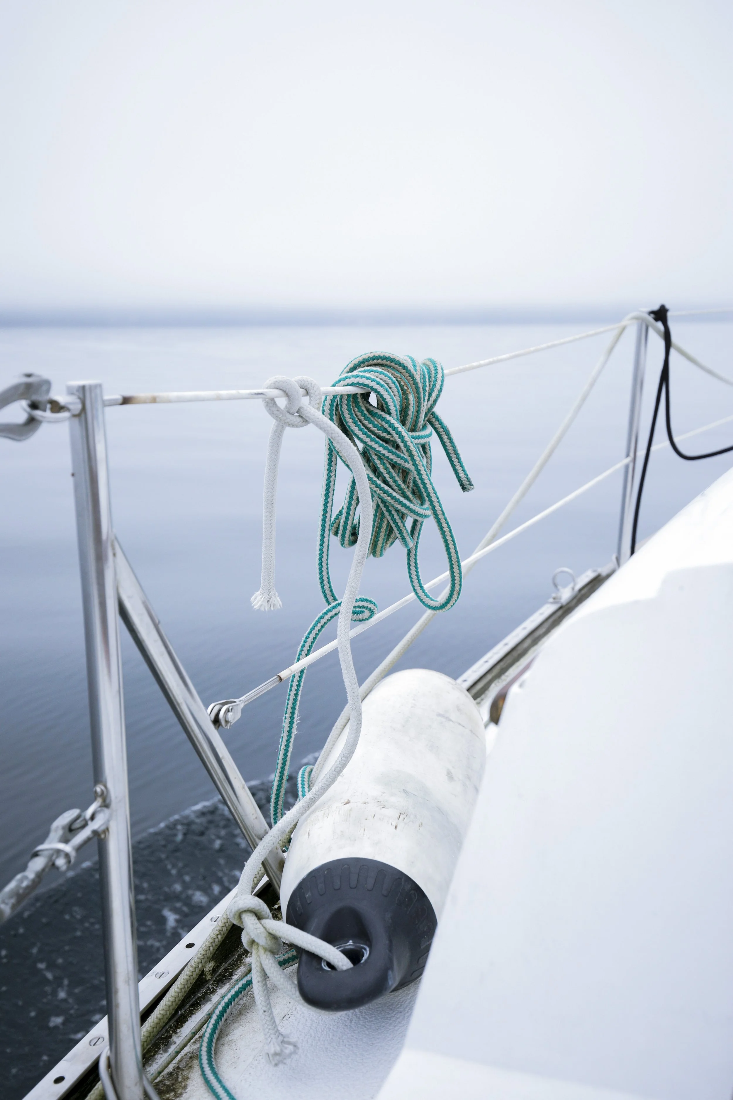 Ropes tied on a boat's railing with a large fender hanging on the side, over calm water with a foggy sky in the background.
