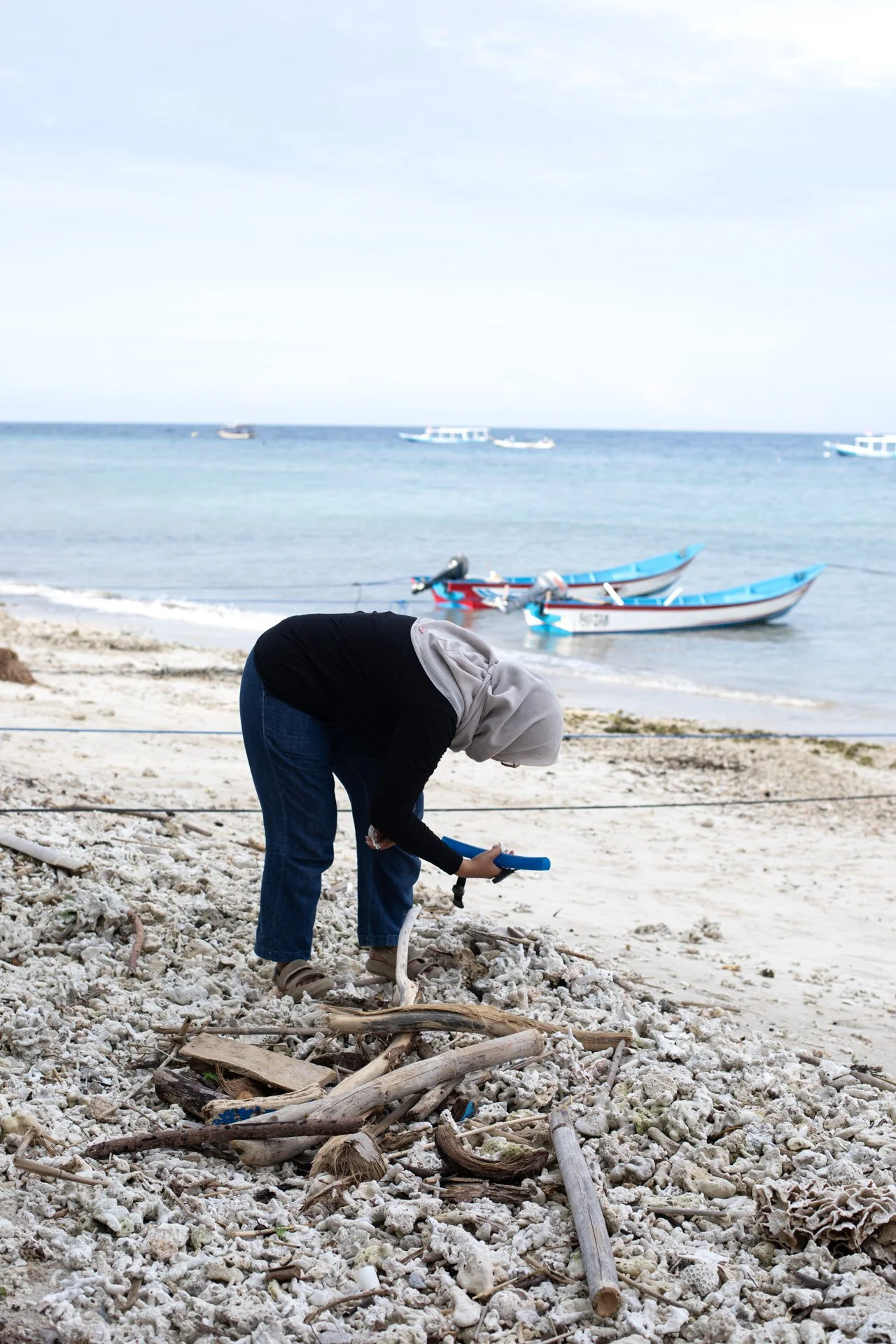 Person in hoodie and jeans bending over collecting sea debris on sandy beach with driftwood, with boats in the water and boats on the horizon