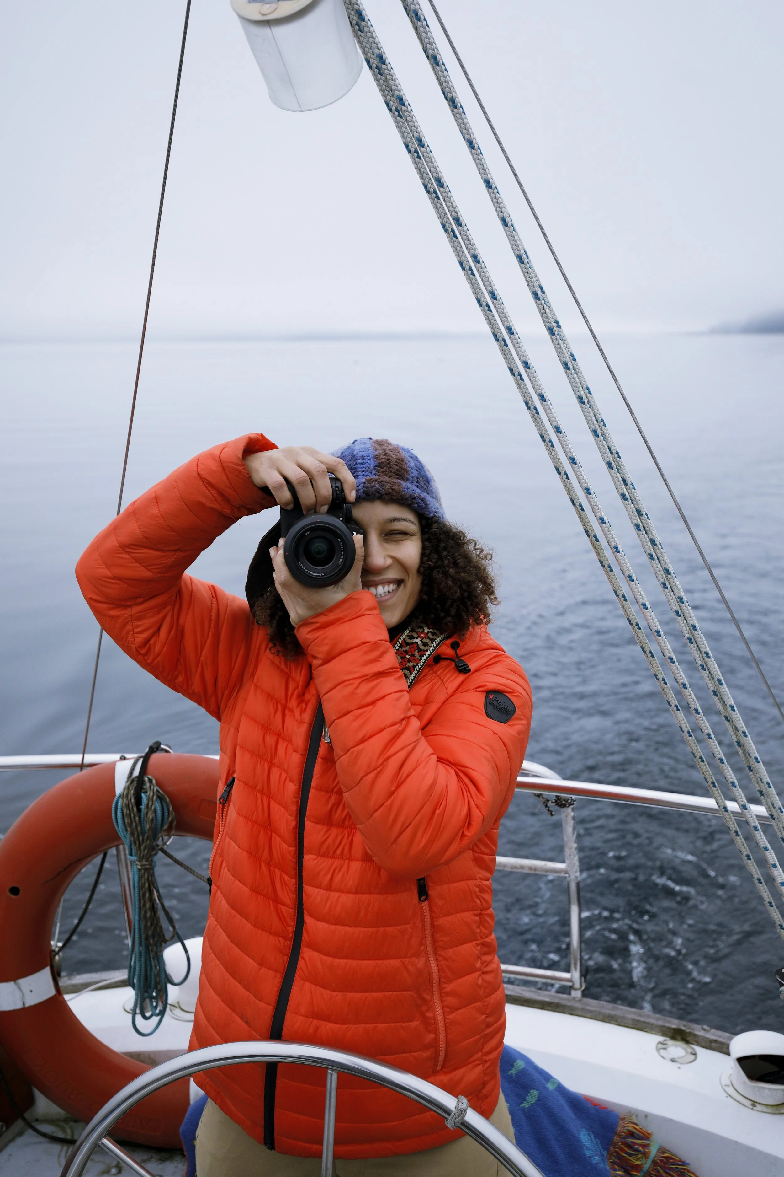 A person with curly hair wearing an orange jacket and a multicolored hat is taking a photo with a camera on a boat, with the water and sky in the background.