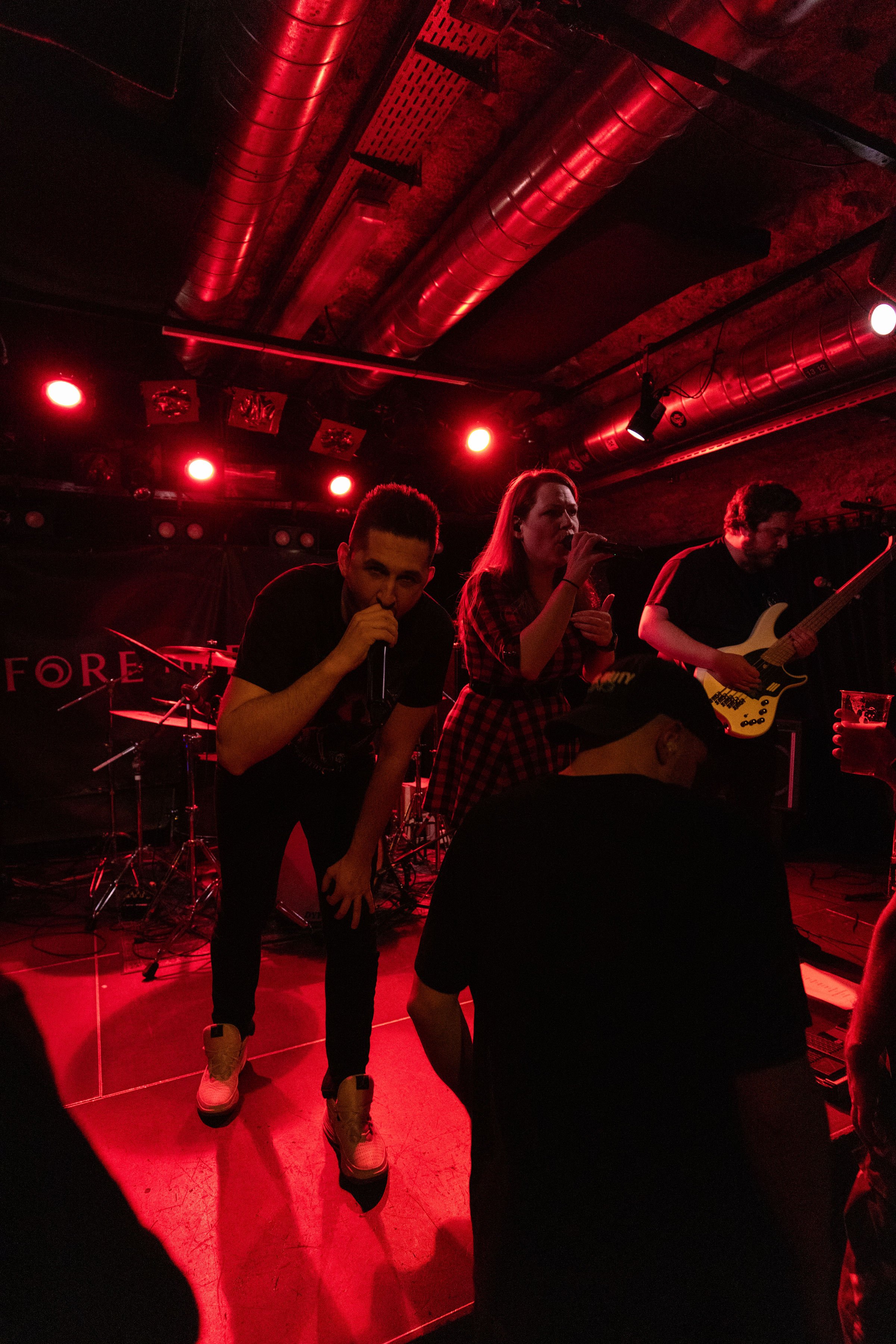 Before The Decay performing on stage with red lighting, two singers with microphones, one man and one woman, and a guitarist in background, audience in front.