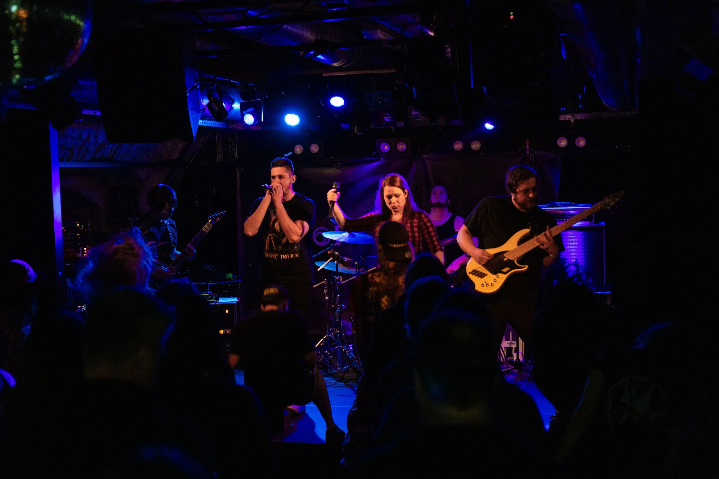 Before The Decay performing live on stage with instruments and microphones, under blue stage lights, with an audience watching.
