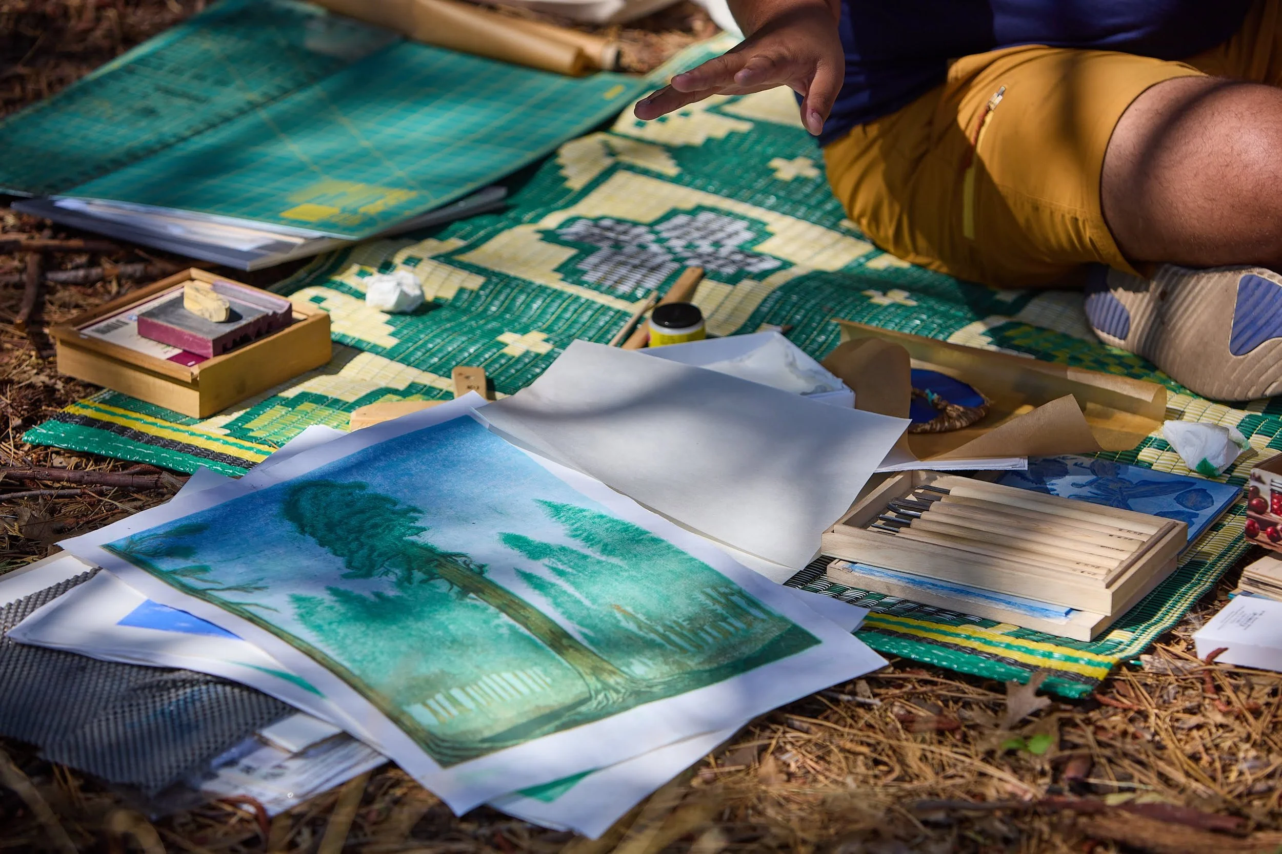 A person sitting outdoors on a colorful mat, surrounded by art supplies, watercolor paintings, and paper.
