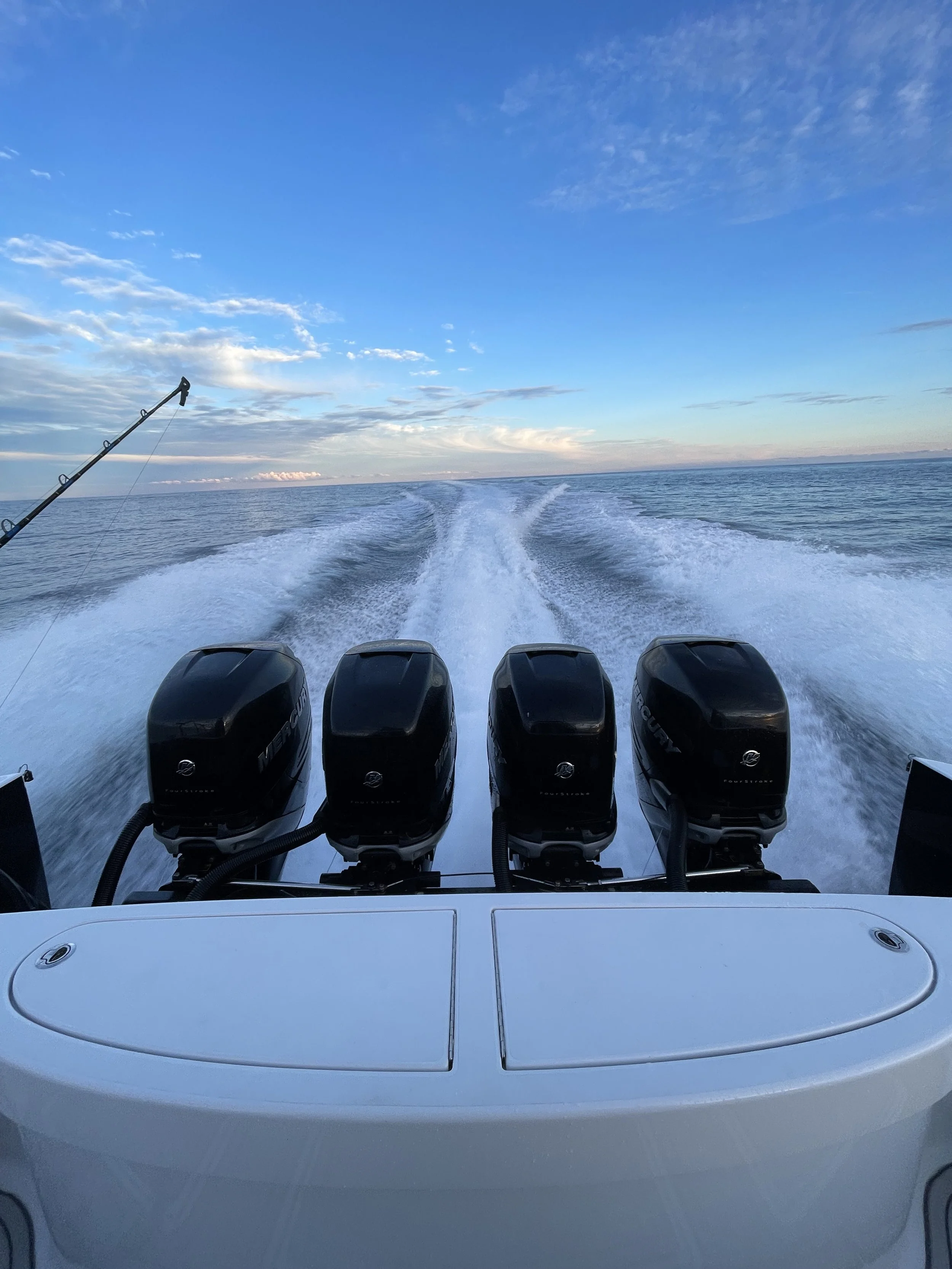Back view of a boat with four outboard motors on a vast body of water, a fishing rod to the left, and a sky with clouds and blue hues.