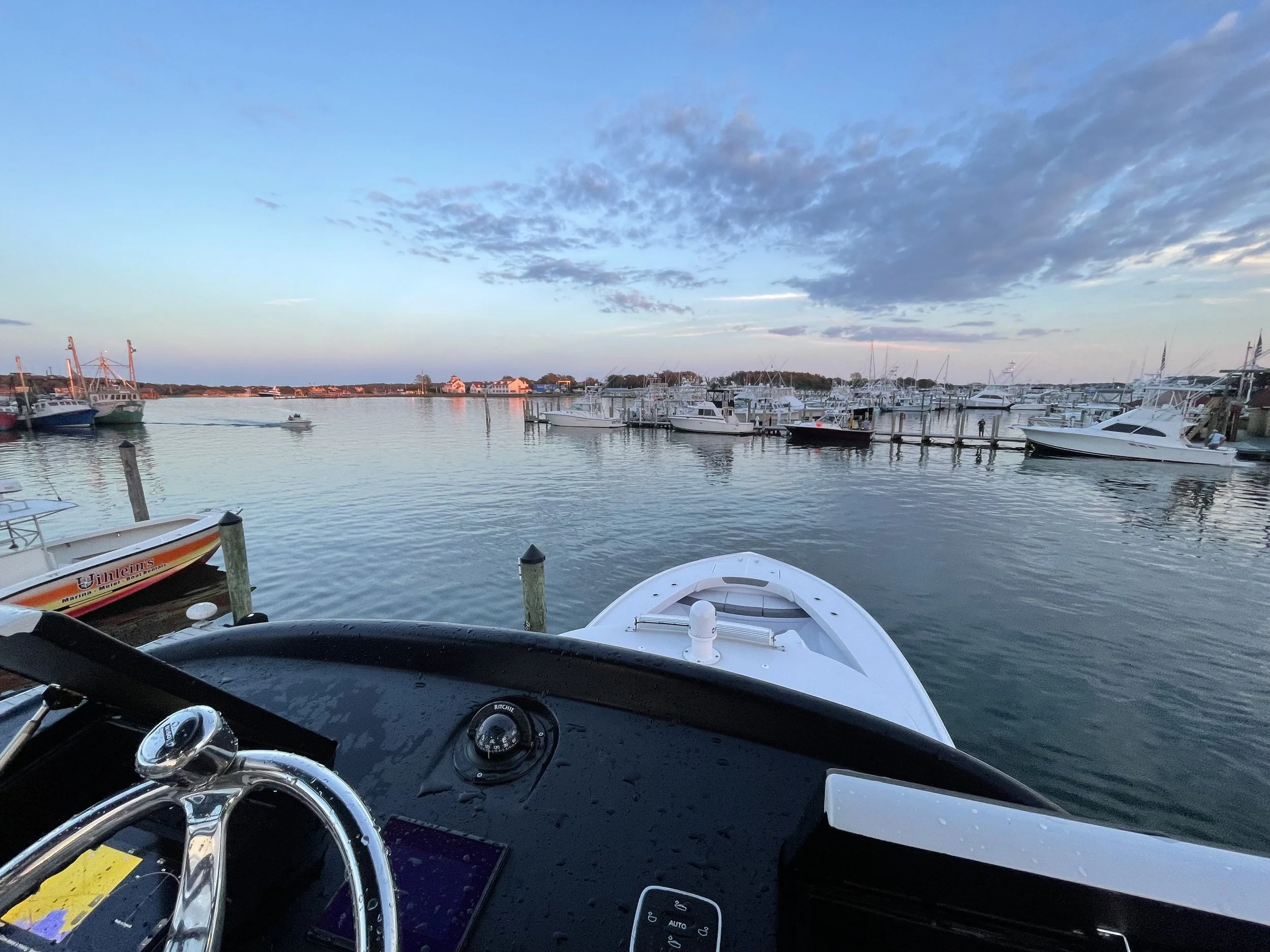 View from a boat dashboard overlooking a marina with various boats and yachts docked, calm water, and a partly cloudy sky at sunset.