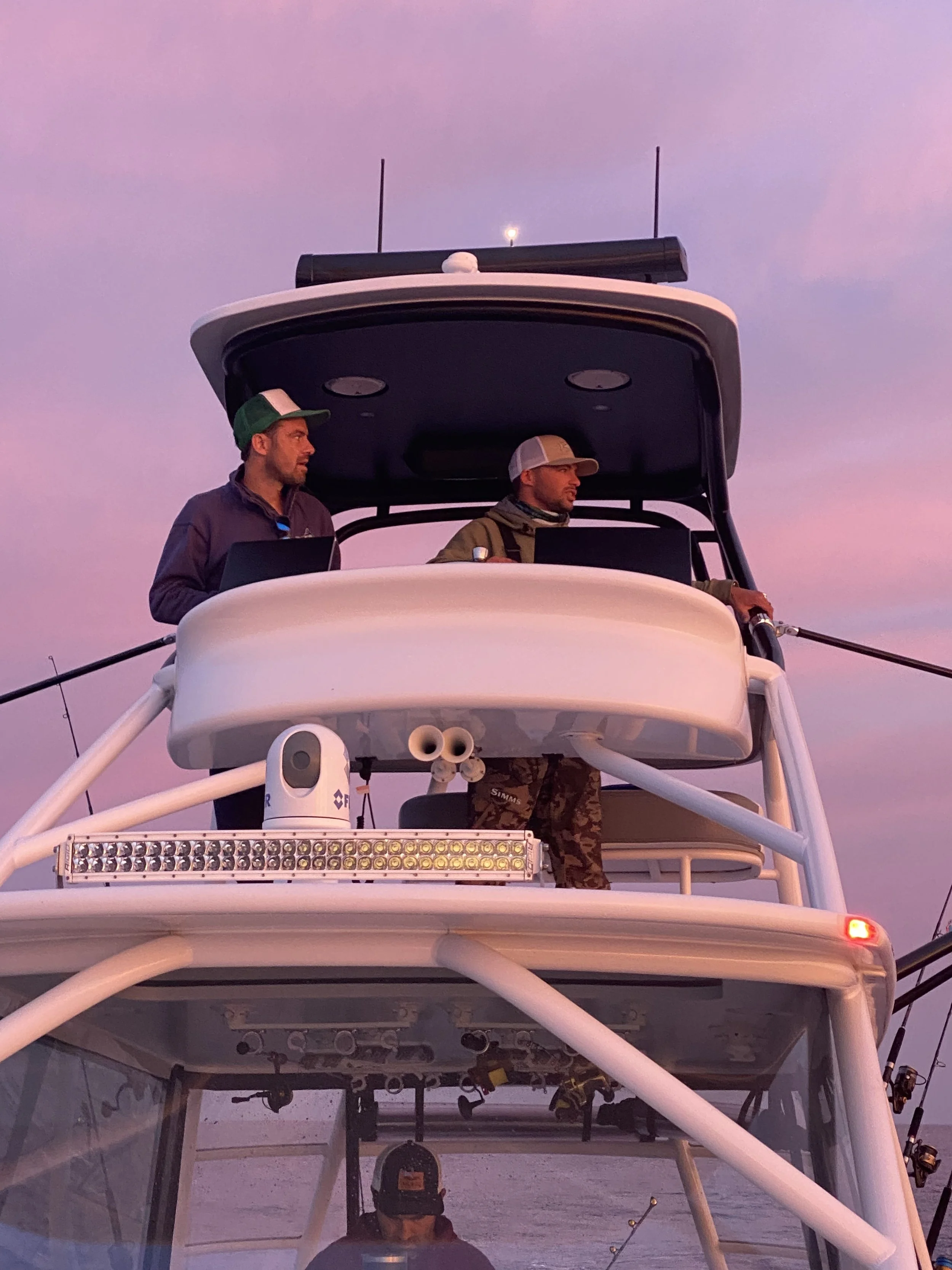 Two men on the top deck of a boat at sunset, with a purple sky. One man is wearing a green and white cap, and the other is wearing a gray and white cap, both looking into the distance.