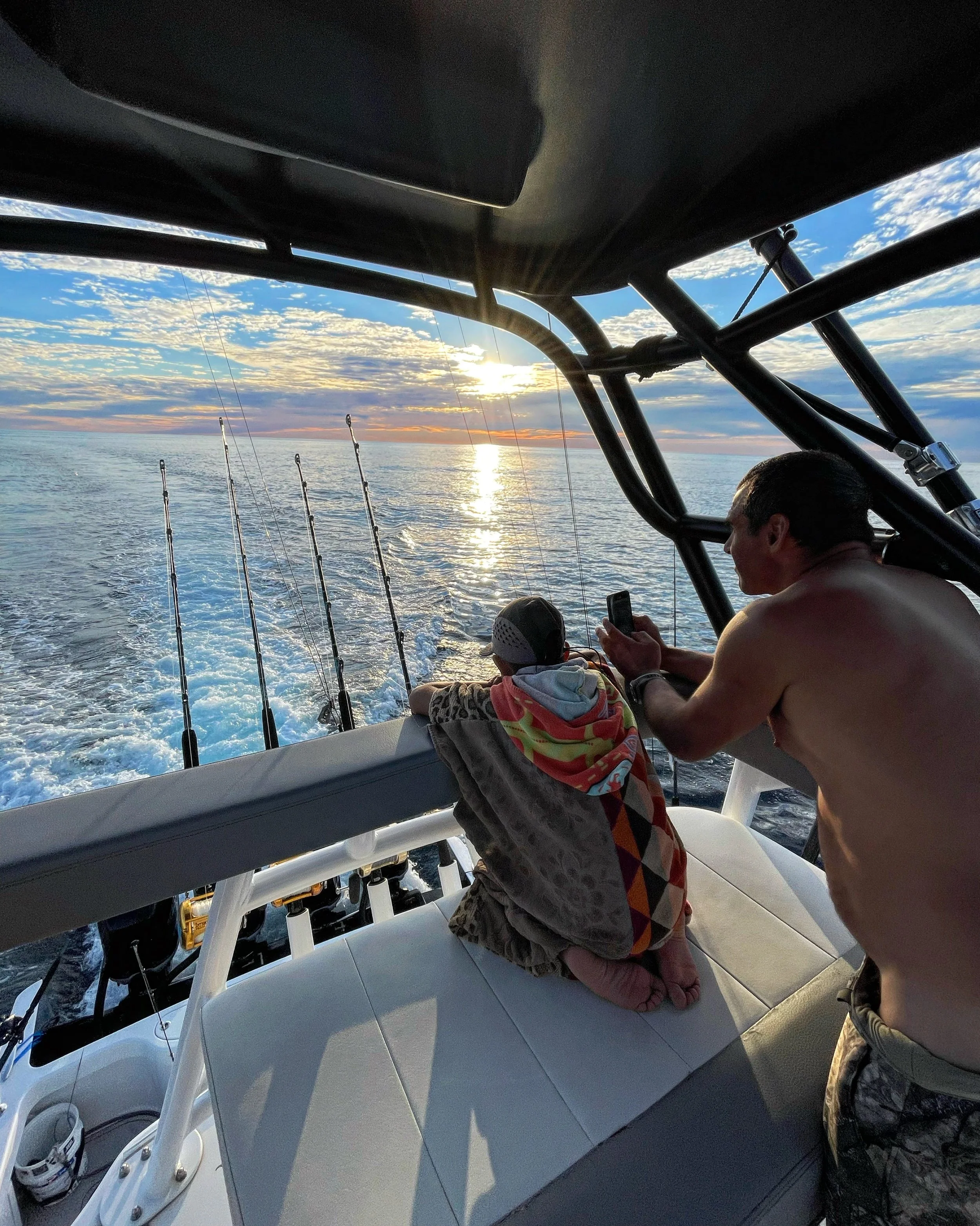 A man and a young boy sitting on a boat watching the sunset over the ocean. The man is shirtless, and the boy is wrapped in a colorful towel. Fishing rods are visible, and the sky is partly cloudy with the sun near the horizon.