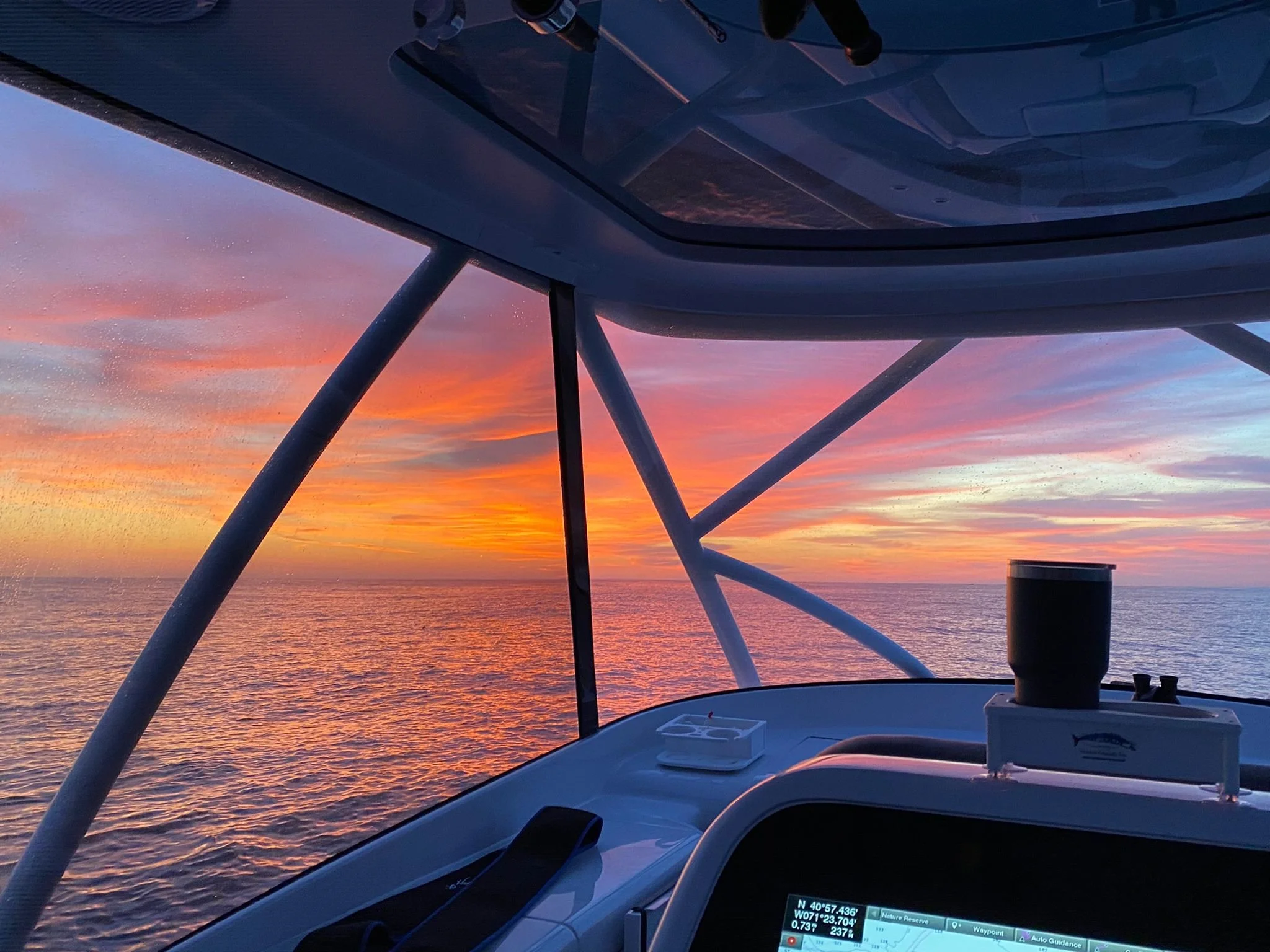 View from a boat at sunset showing a colorful sky with shades of orange, pink, purple, and blue over the ocean. Part of the boat's structure and dashboard are visible in the foreground.